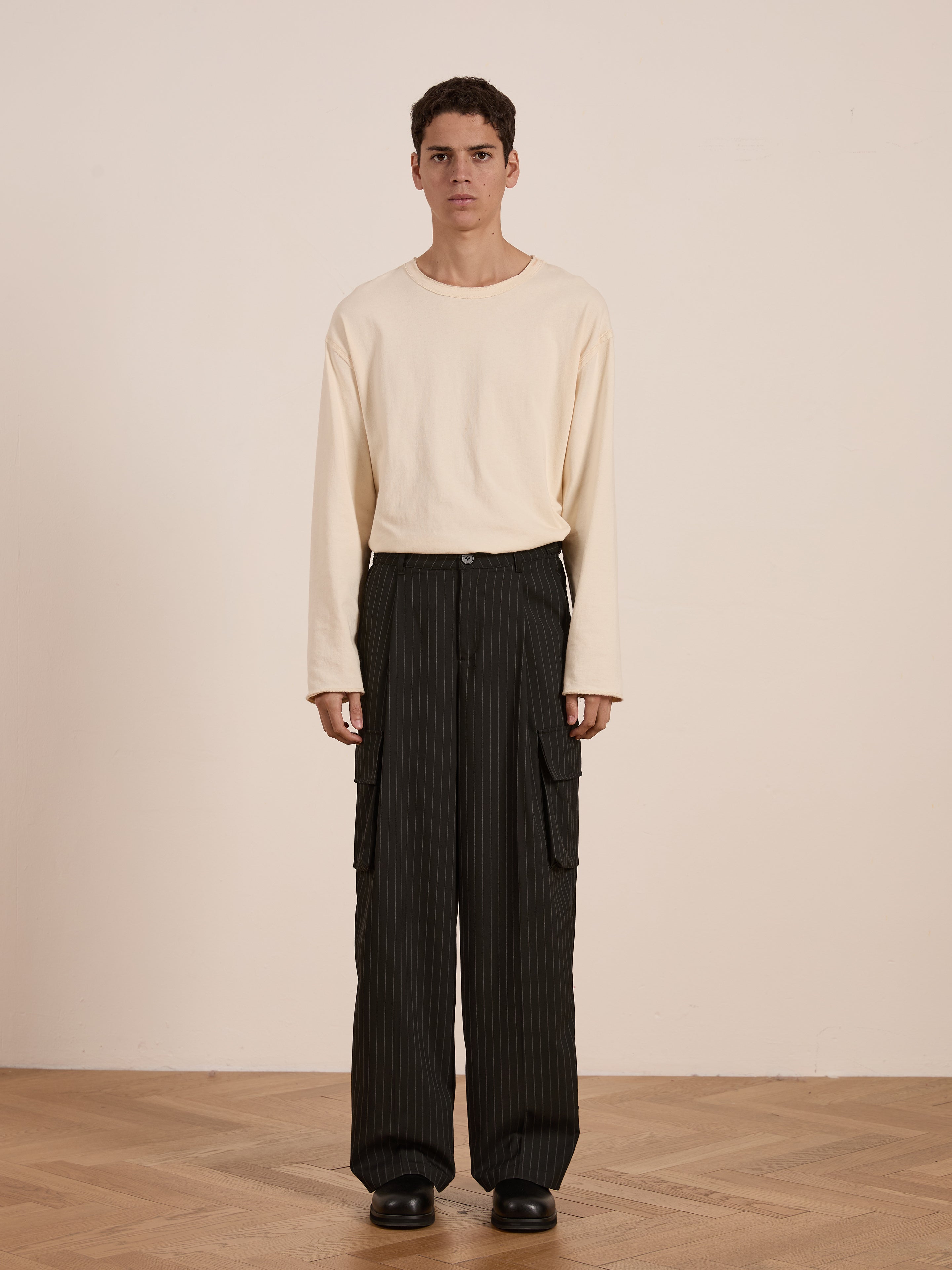 Against a plain background, a person stands wearing a long-sleeve beige shirt and FOUND's Hava Pinstripe Pleated Cargo Trousers with adjustable side tabs. Their sleek black shoes complete the look.