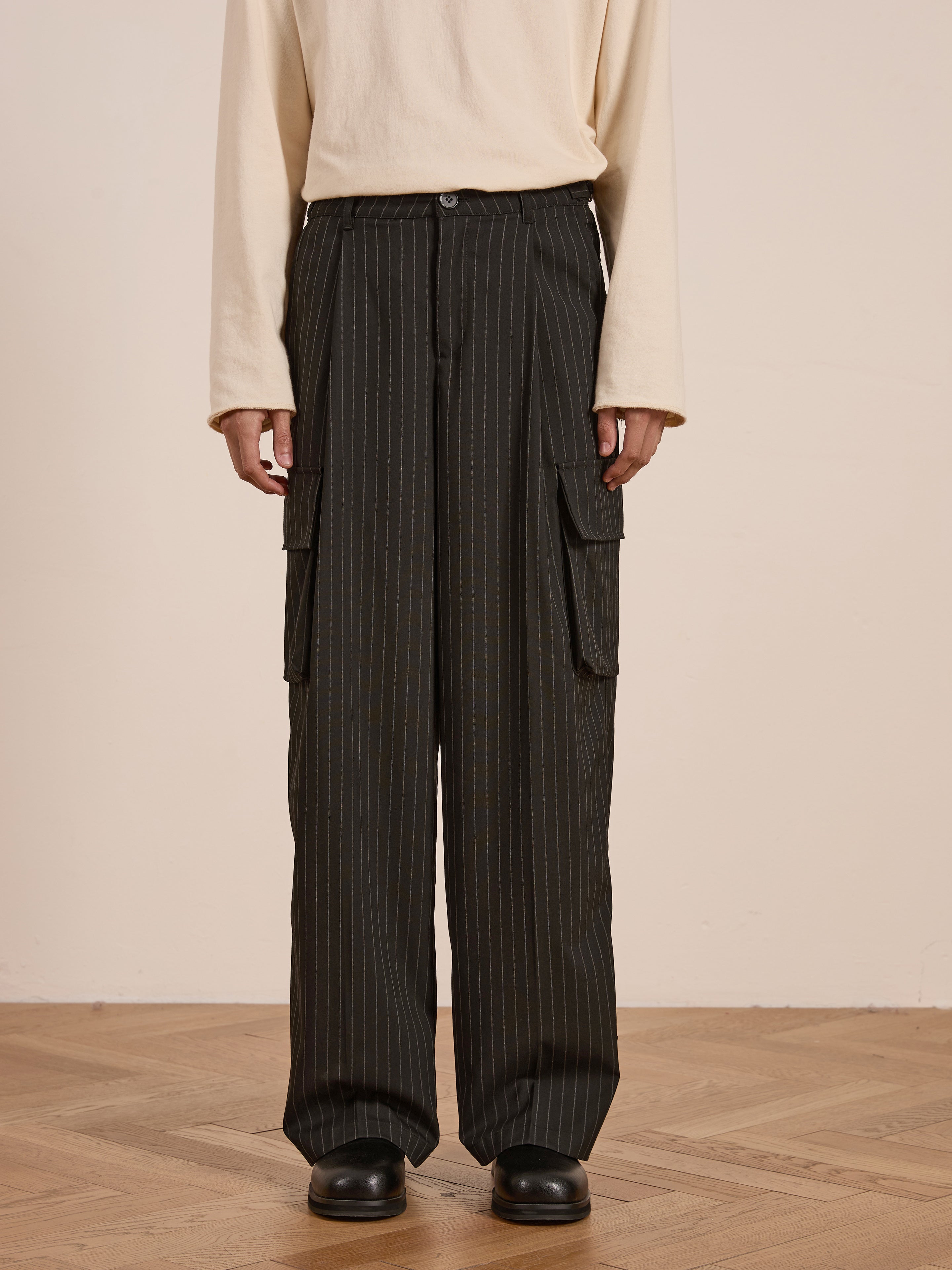 A person in a cream long-sleeve shirt and Hava Pinstripe Pleated Cargo Trousers by FOUND, made from a cotton-blend, stands on a wooden floor wearing black boots for the ideal utility look.