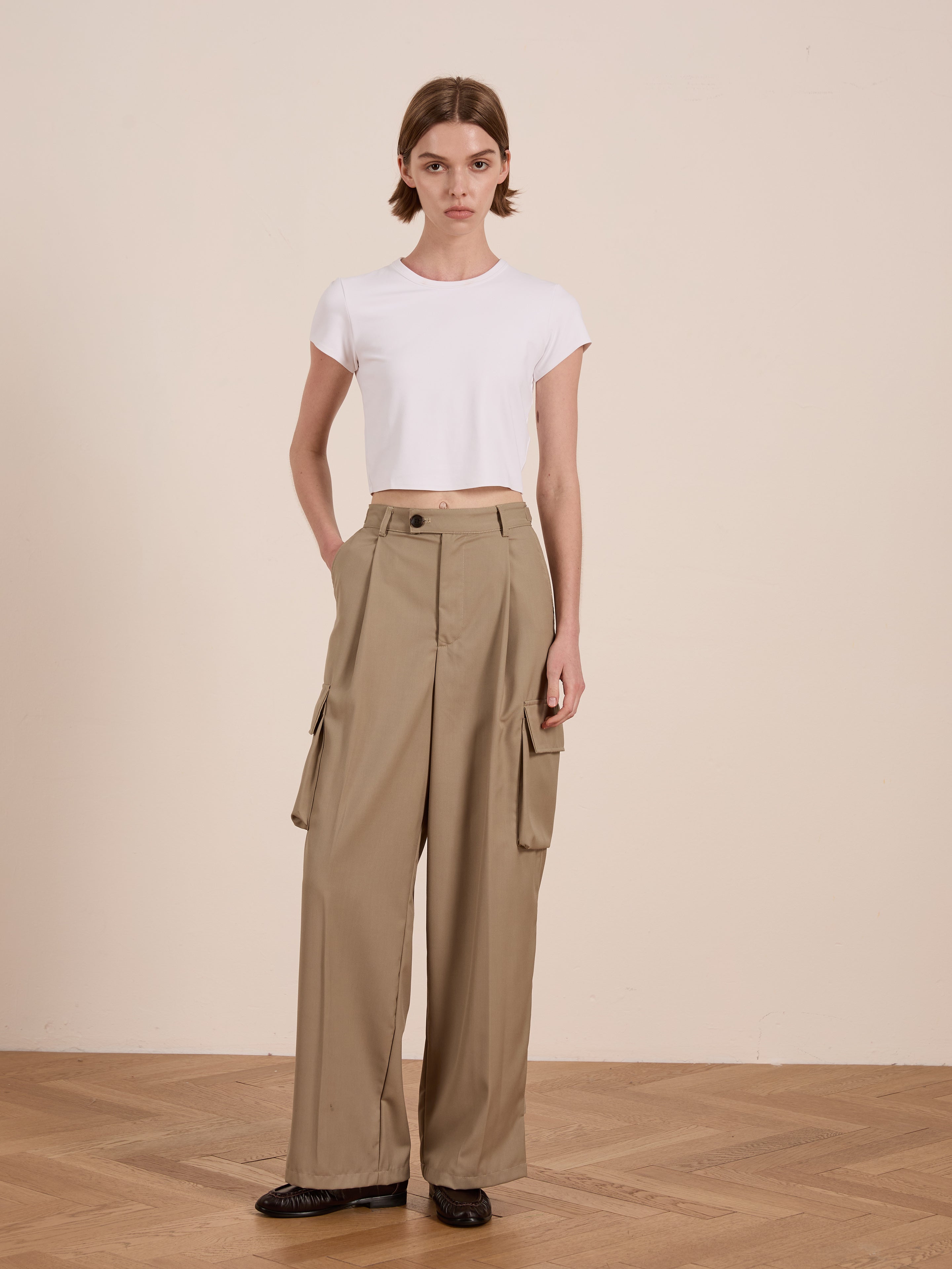 A person in a white cotton blend crop top and Hava Pleated Cargo Trousers by FOUND stands on a wooden floor against a plain background, embodying utilitarian style.