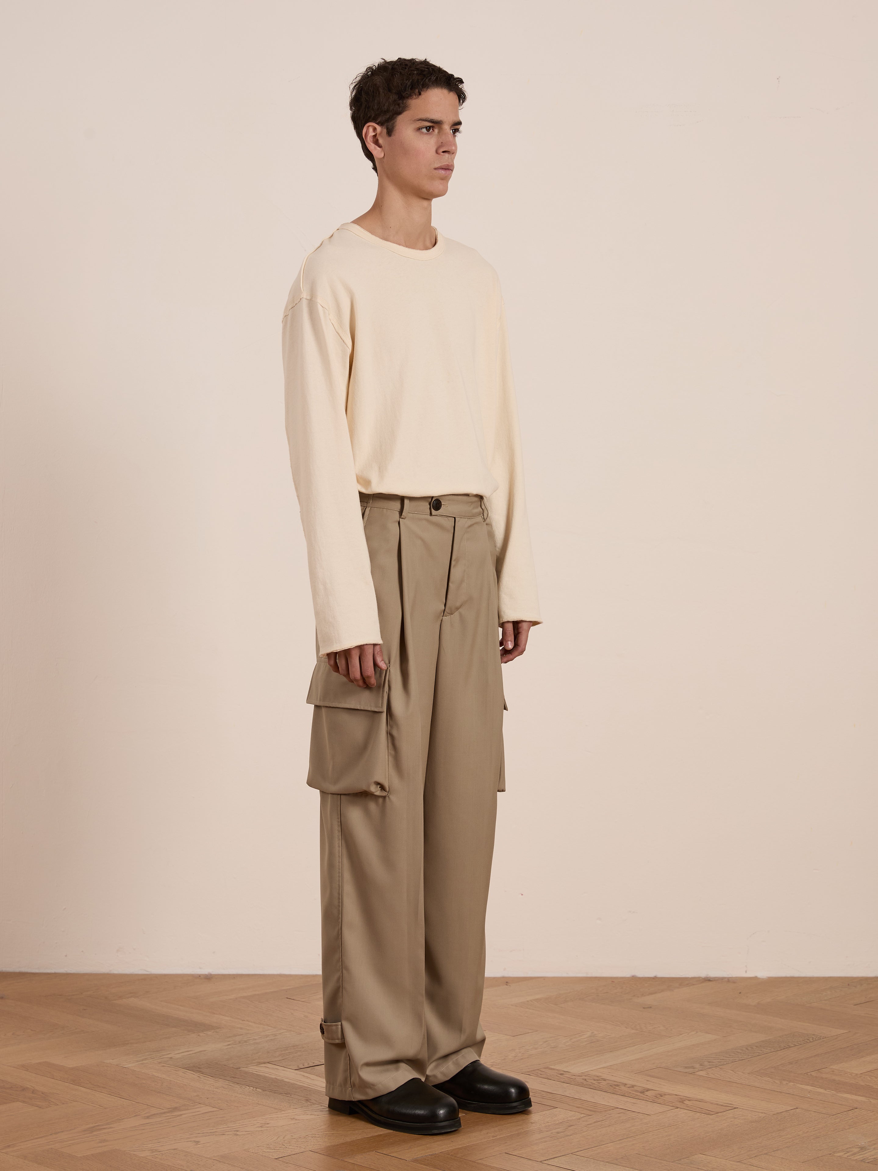 Someone dressed in a cream long-sleeve shirt and FOUND's Hava Pleated Cargo Trousers made from cotton twill stands on a wooden floor against a neutral backdrop.