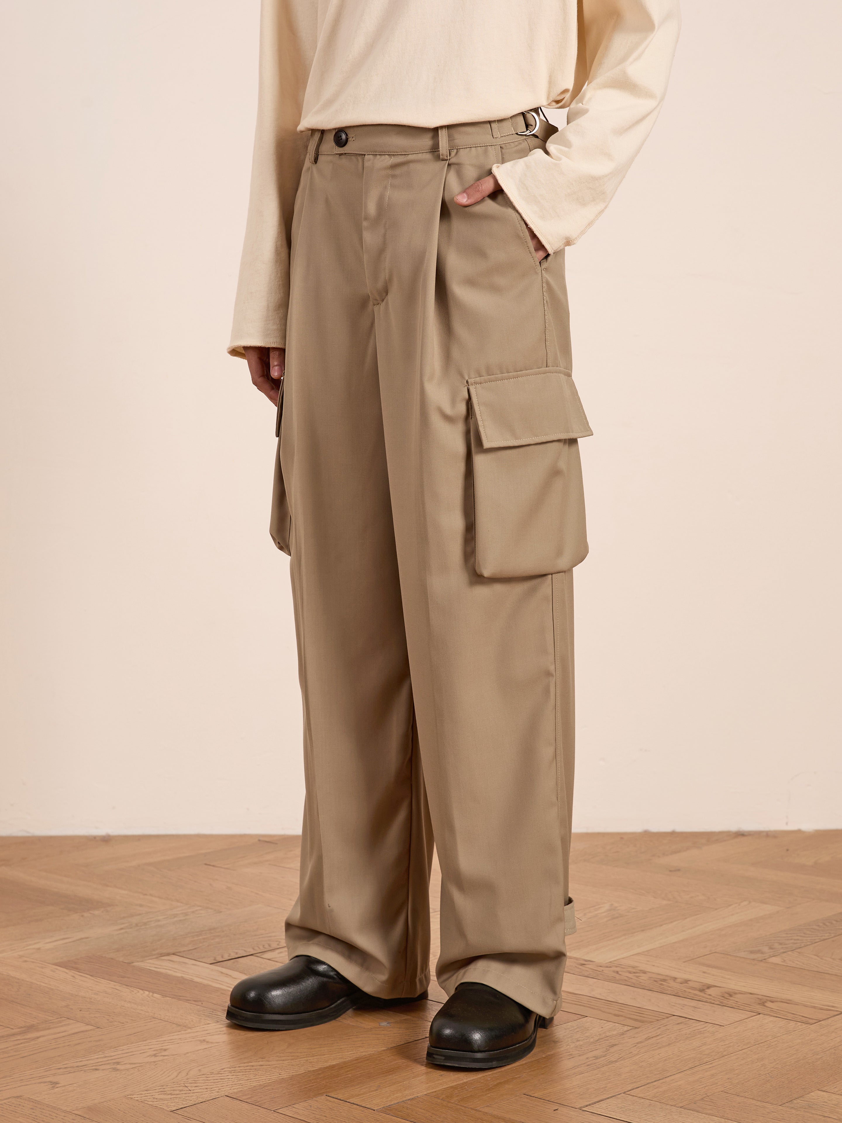 Dressed in FOUND's Hava Pleated Cargo Trousers on a wooden floor, the person wears a long-sleeve cream top, epitomizing the utilitarian style with their cotton blend ensemble.