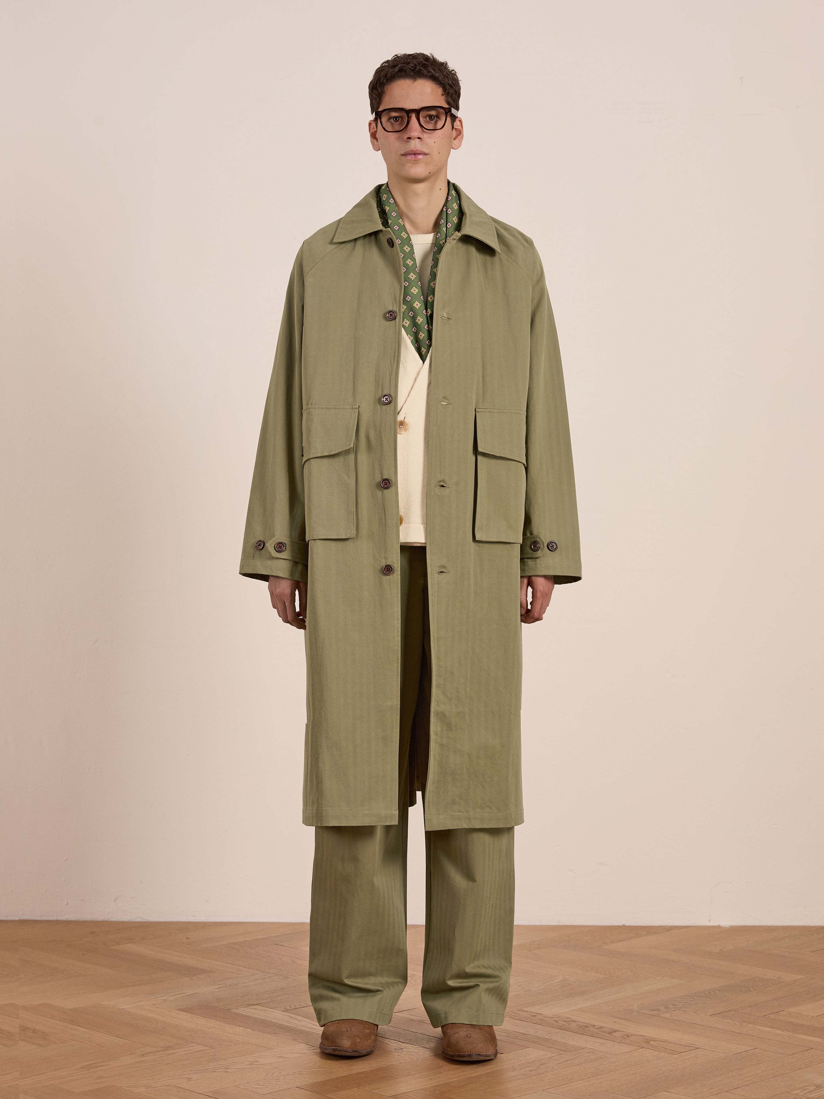 Against a plain light backdrop, someone stands on a wooden floor wearing glasses, brown shoes, matching pants, a green patterned scarf, and FOUND's Herringbone Twill Trench Coat with utility pockets.