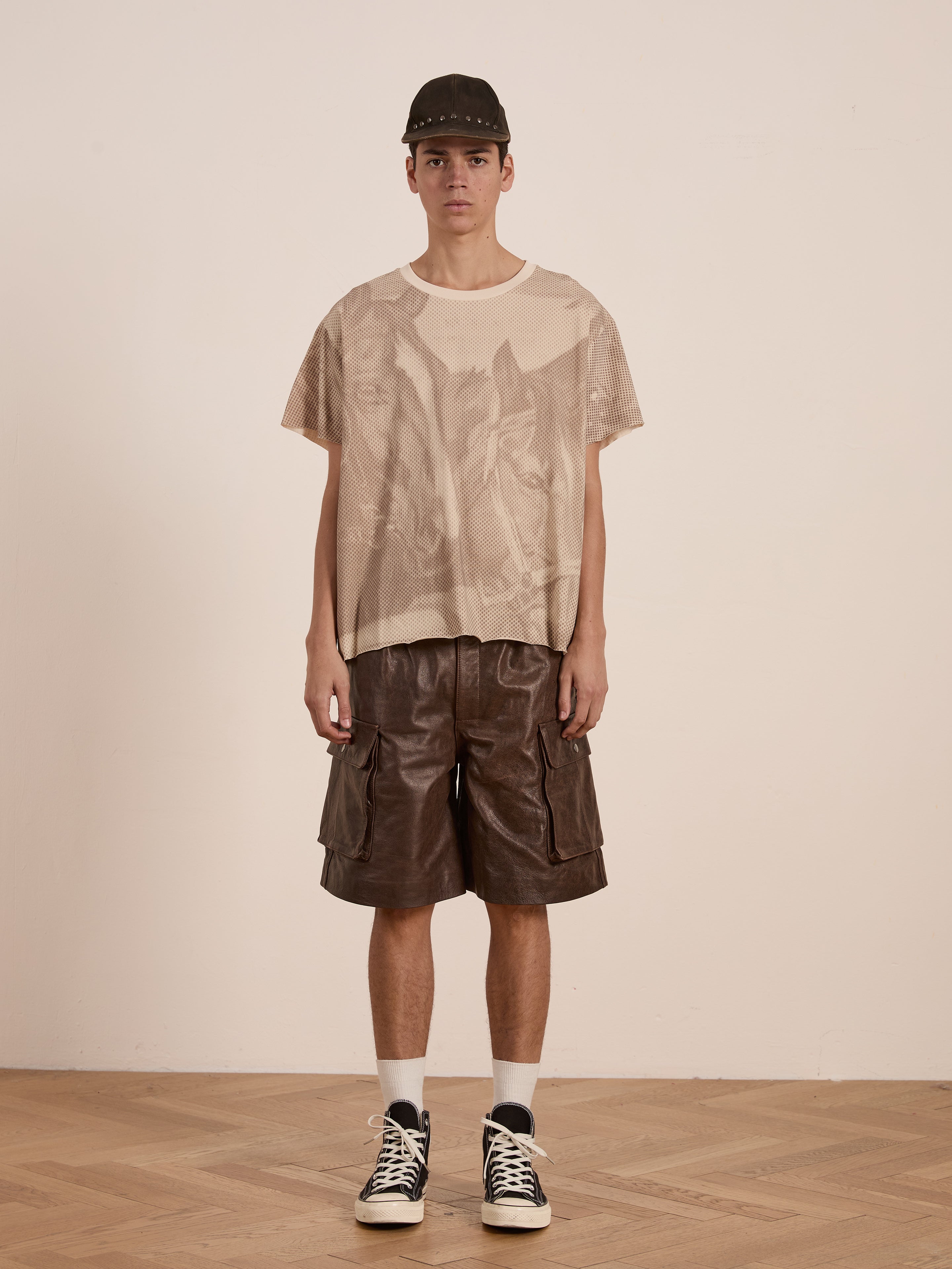 A person indoors sports a relaxed fit FOUND Horse Racer Tee in beige tones, paired with brown cargo shorts, white socks, and black sneakers, set against a plain background.