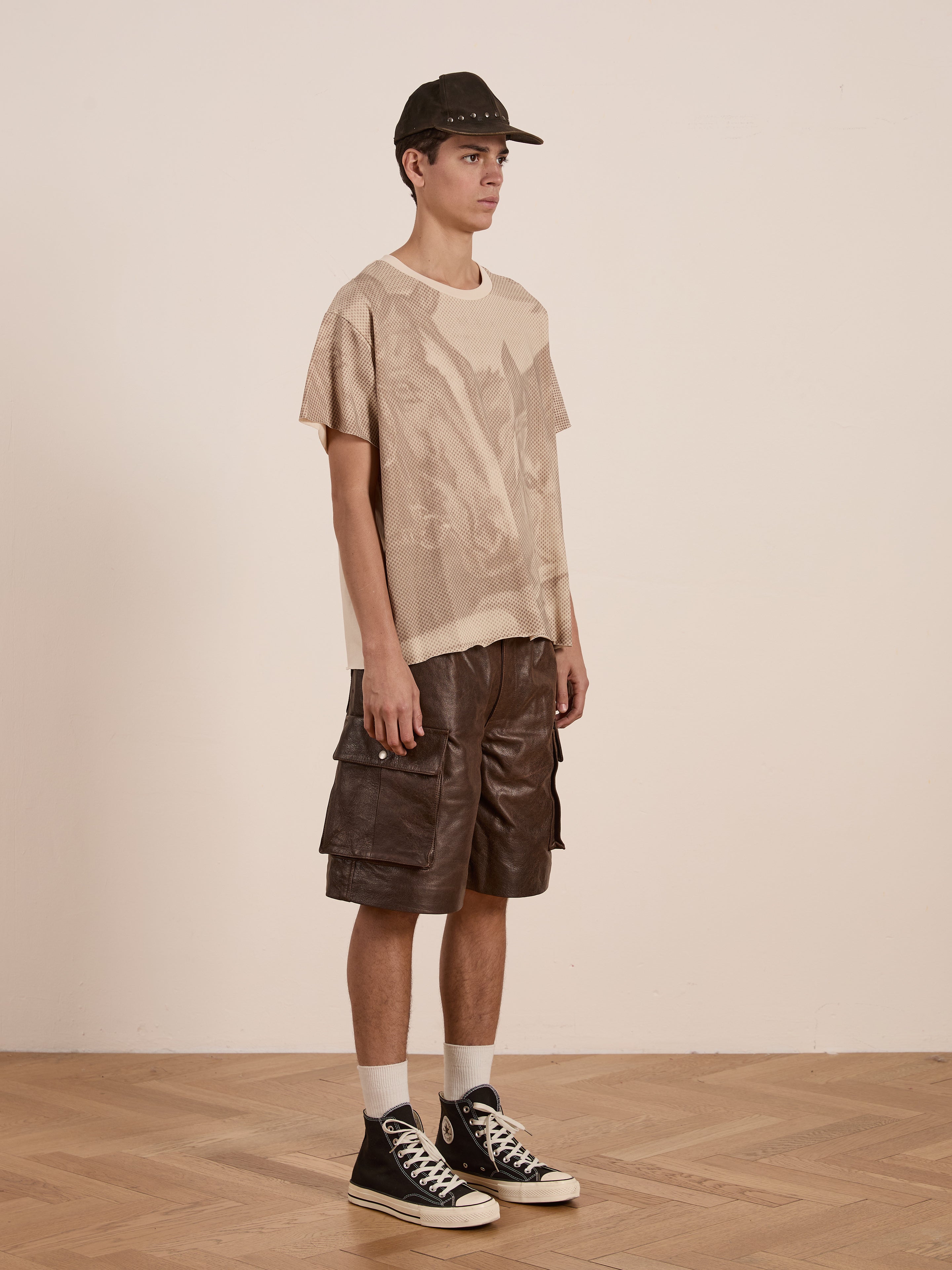 A person stands in profile on a wooden floor, wearing a relaxed-fit beige Horse Racer Tee from FOUND, paired with brown cargo shorts, black sneakers, white socks, and a dark baseball cap against a plain background.