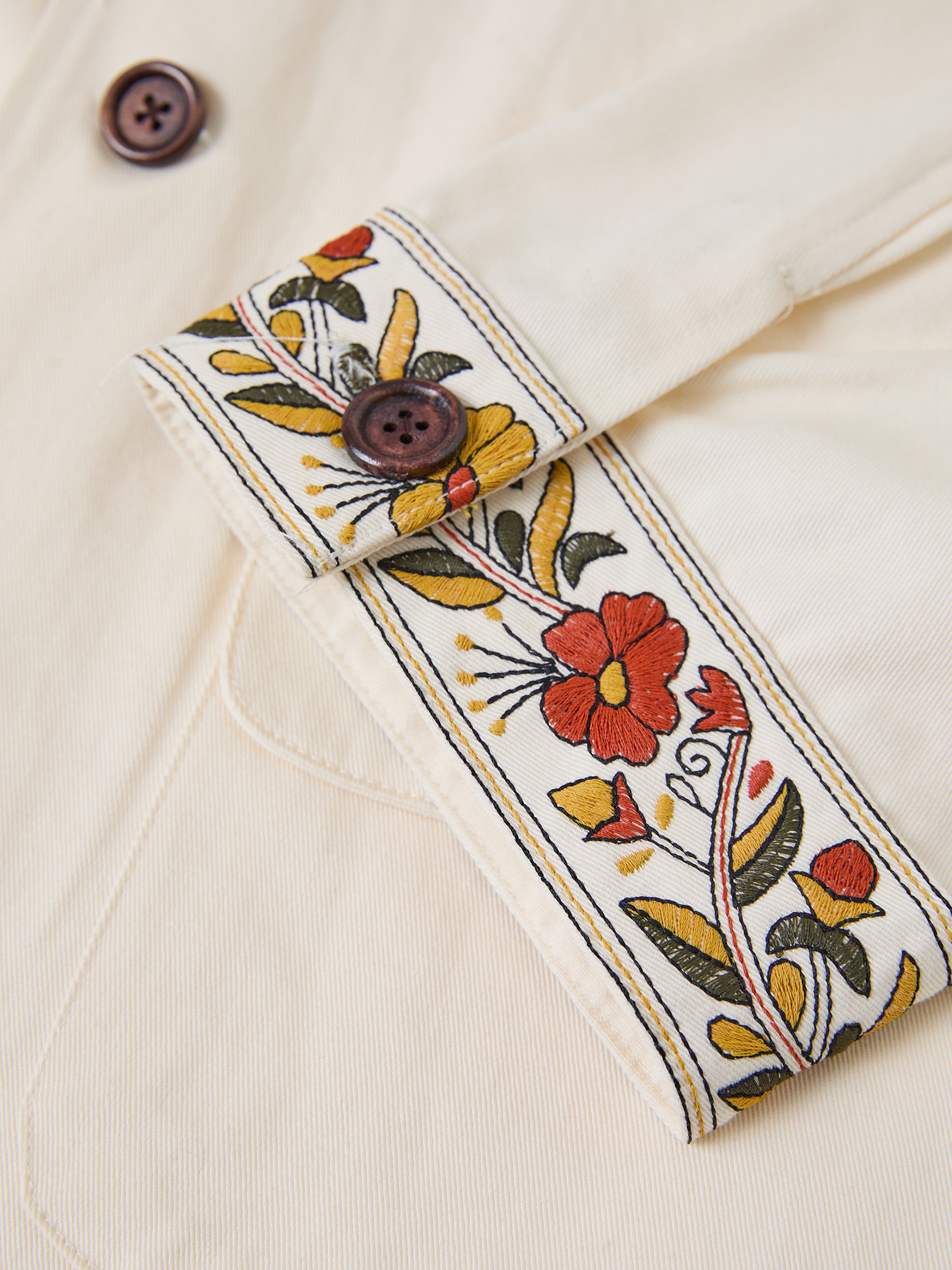 The FOUND Kantha Floral Work Jacket features a vintage charm with its close-up of beige fabric, embroidered floral patterns, and brown buttons. The intricate red, yellow, and green designs enhance the textured background beautifully.