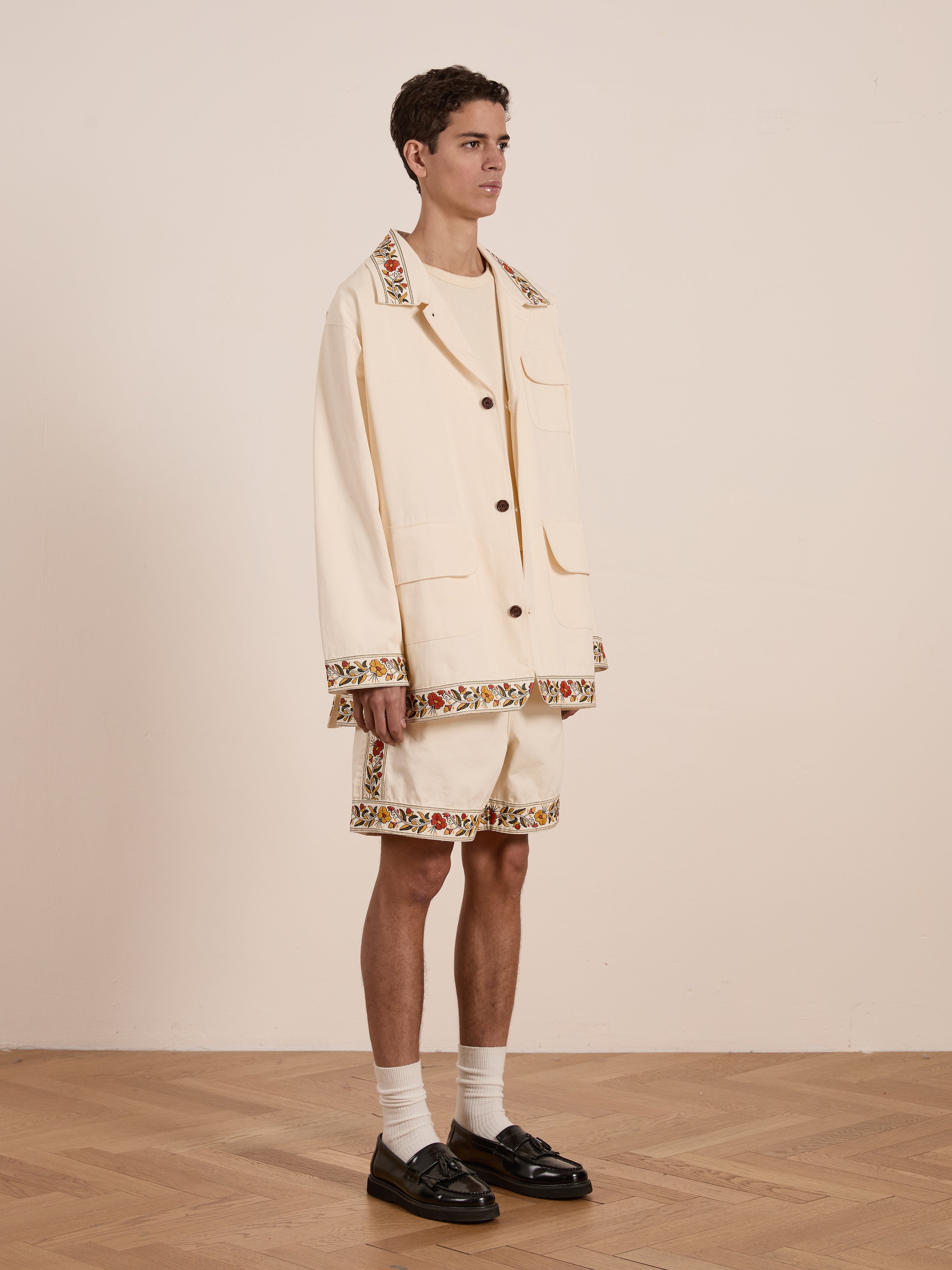 A person models an oversized beige Kantha Floral Work Jacket by FOUND with shorts featuring floral trim, paired with black loafers and white socks, exuding a vintage vibe against a plain backdrop.