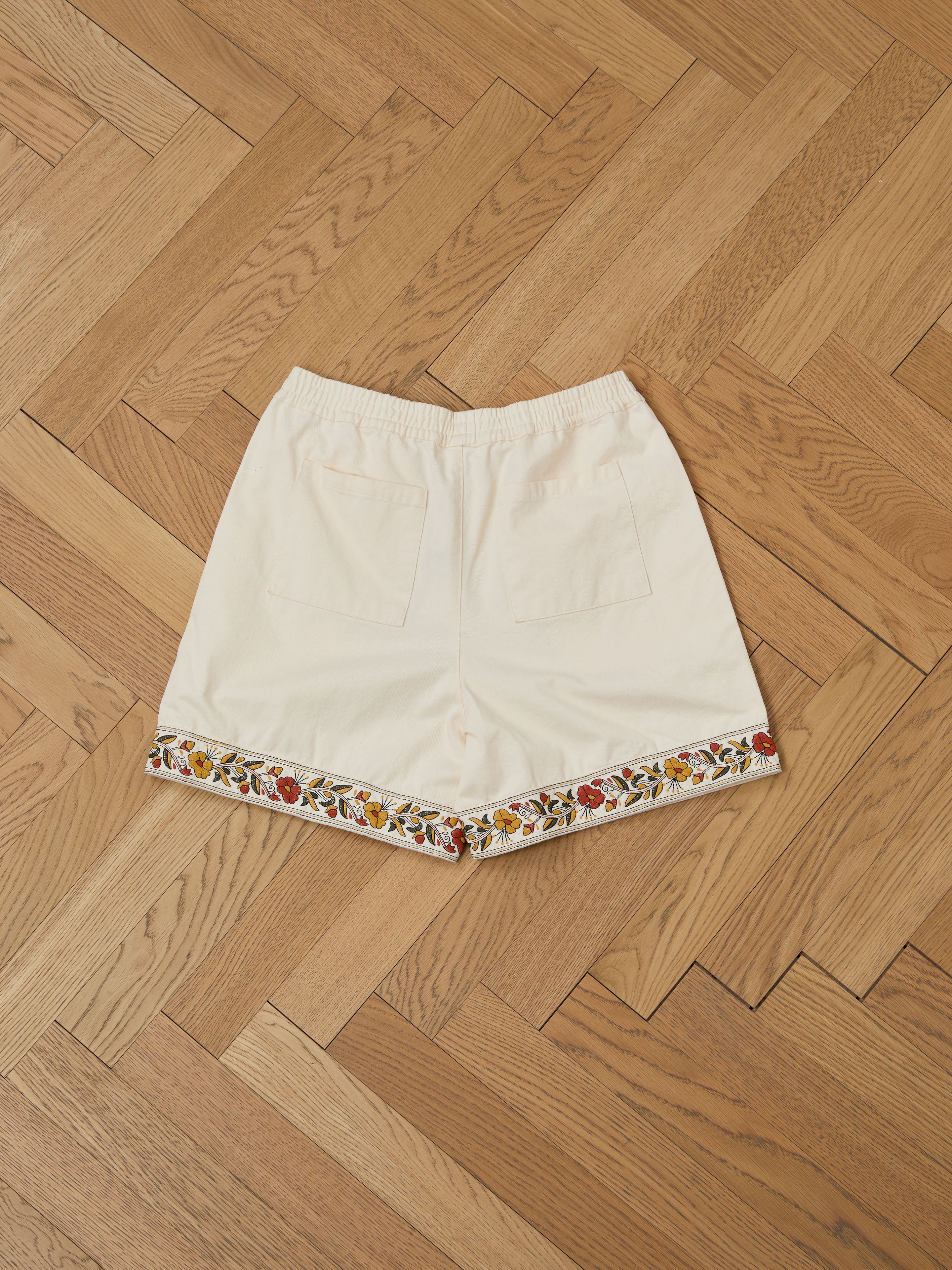 FOUND's Kantha Floral Work Shorts, made of 100% cotton, rest on a wooden herringbone floor. These versatile beige shorts feature two back pockets and delicate floral embroidery on the hem for an elegant touch.