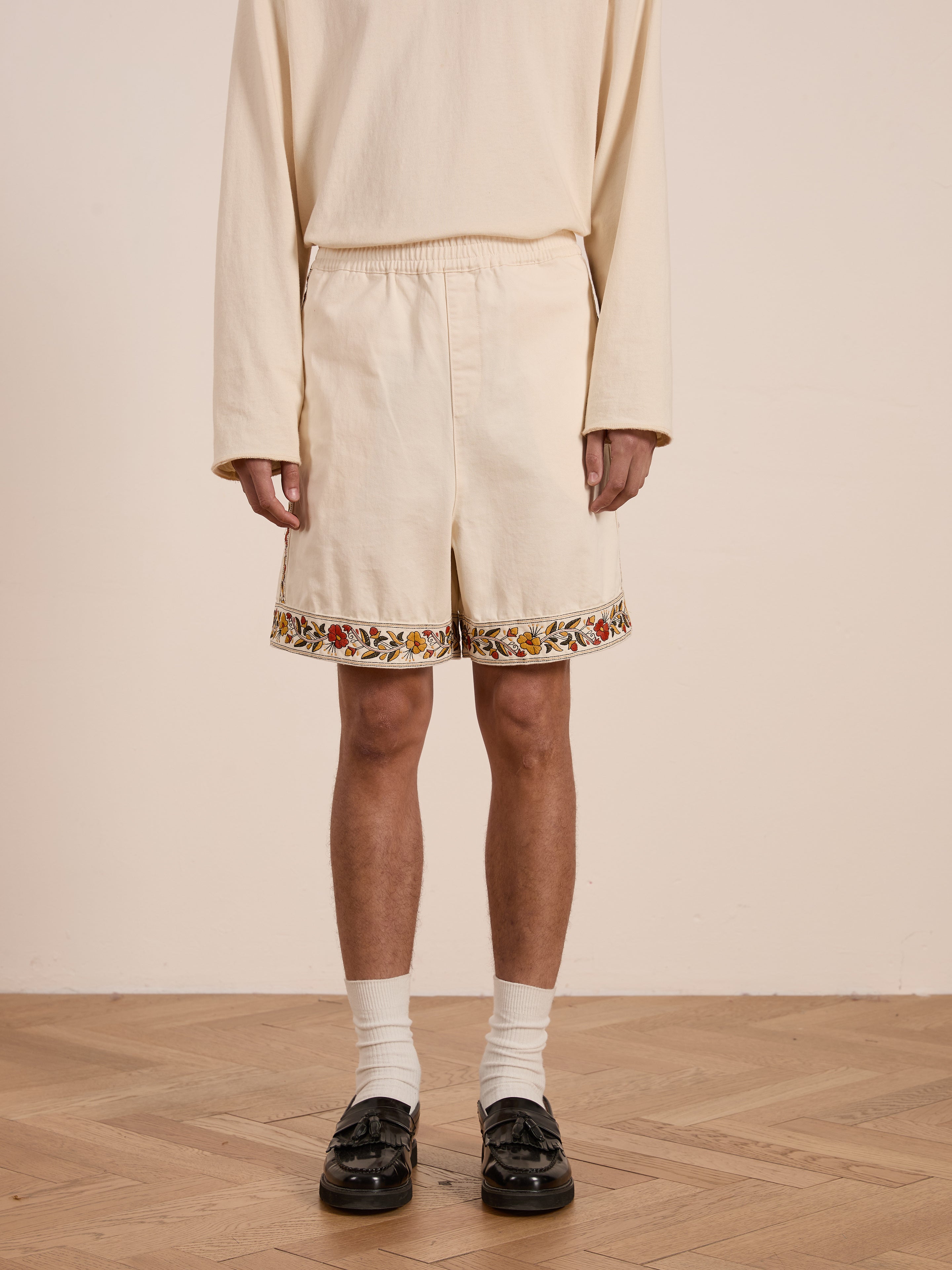 A person wearing a beige long-sleeve shirt and Found's Kantha Floral Work Shorts, both featuring delicate floral embroidery, along with white socks and black shoes, stands on a wooden floor.