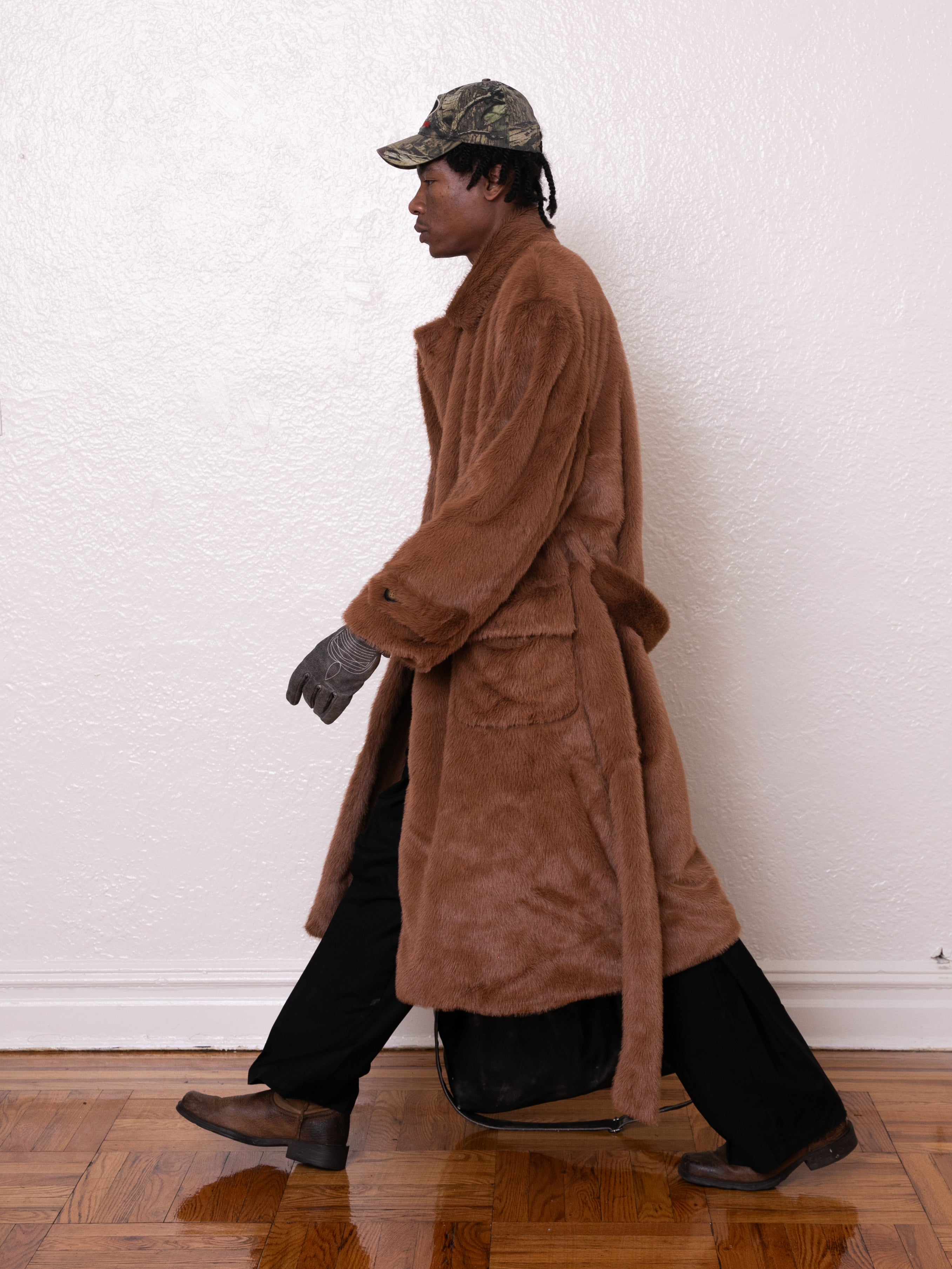 A person strides in profile, wearing the oversized Long Faux Fur Coat from FOUND with a trench shape. Paired with black pants, gray gloves, and a camo cap, they stand against a plain white wall and wooden floor.