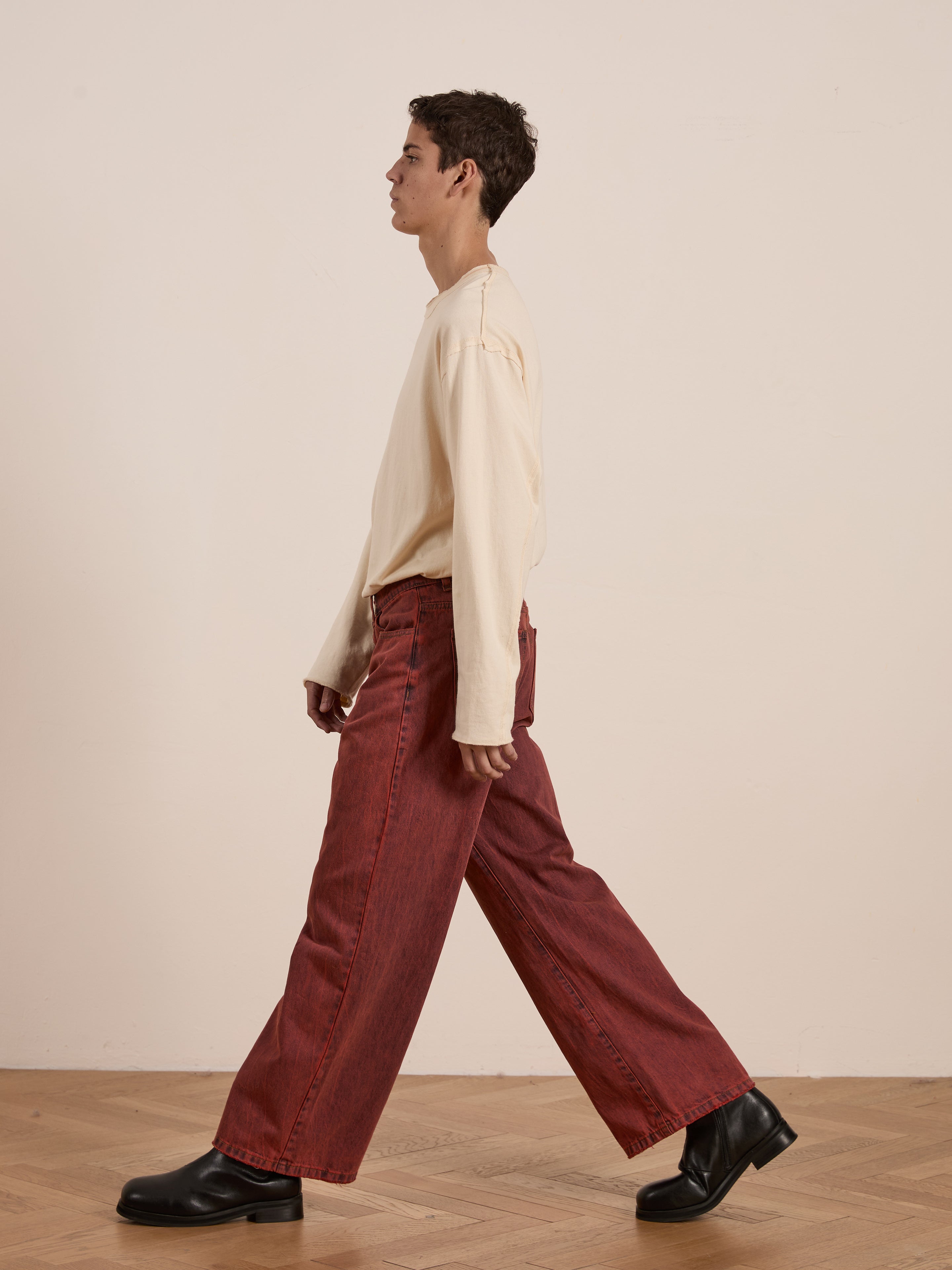 A person in a beige long-sleeve top and FOUND's Lacy Baggy Jeans in vintage red wash walks on a wooden floor against a plain background.