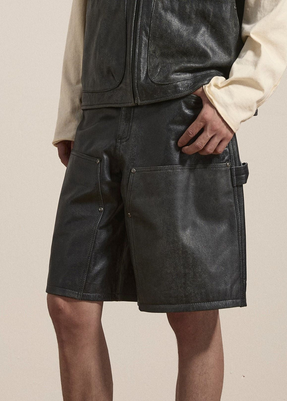 A person wearing a black leather vest and Found's Distressed Genuine Leather Carpenter Shorts, which feature large front pockets and a side loop. These stylish shorts are made from ethically sourced materials and boast a double-knee silhouette for added durability.