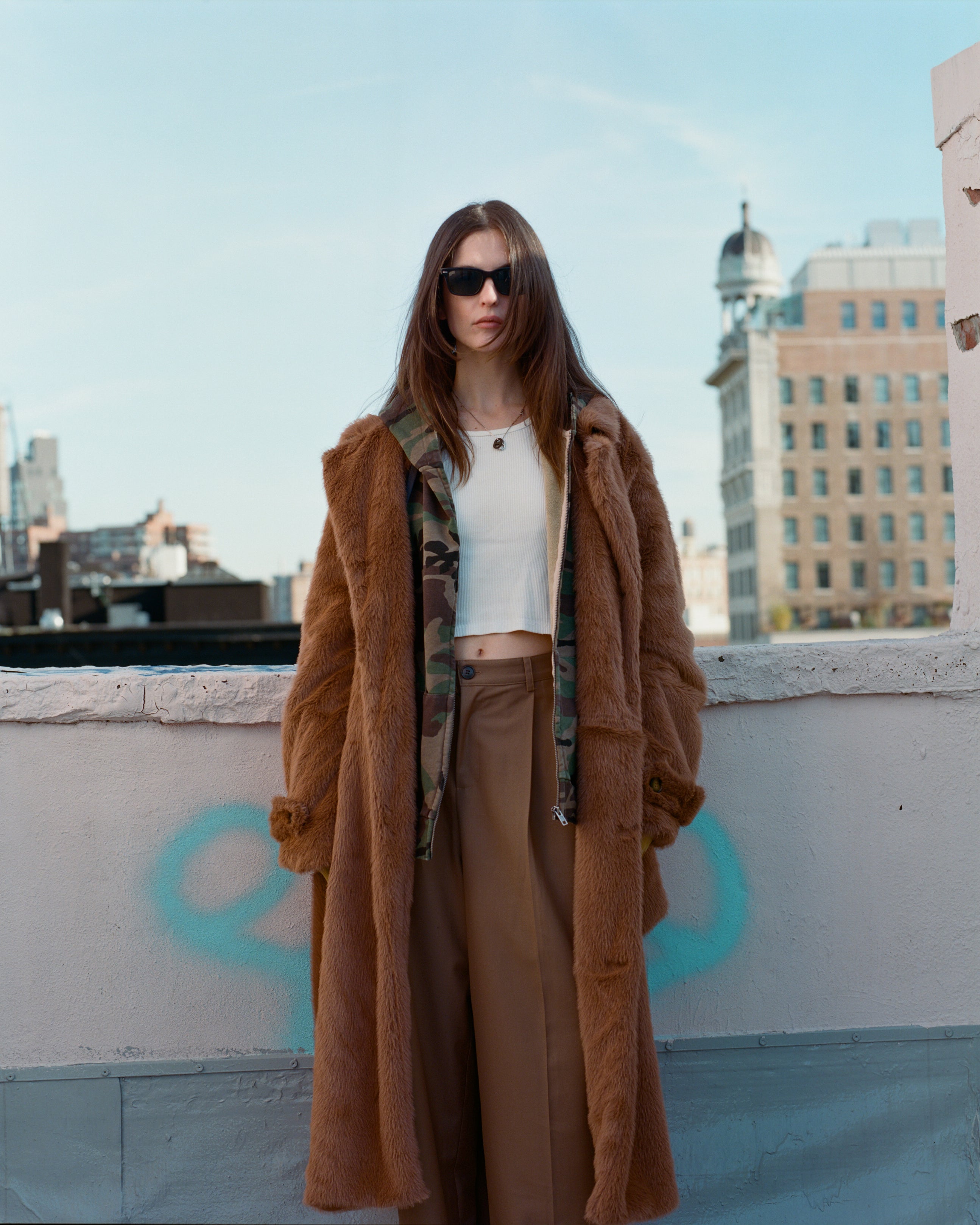 A person wearing the Long Faux Fur Coat by FOUND, along with a white shirt and sunglasses, stands on a rooftop with city buildings in the background.