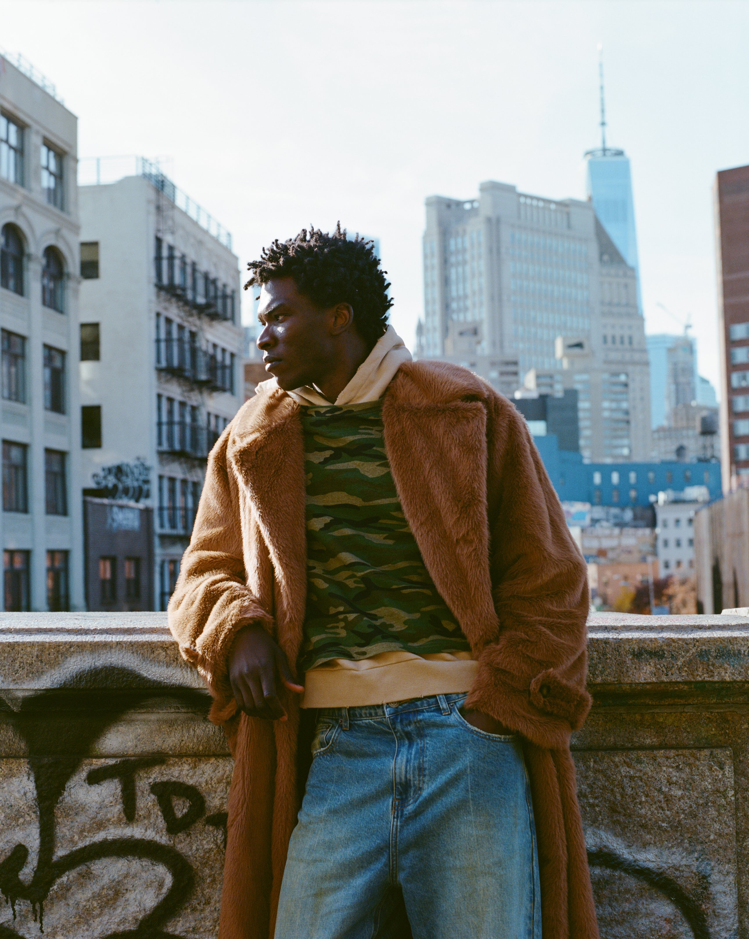 A person in a long faux fur coat by FOUND and jeans leans against a graffiti-covered wall, with city buildings in the background.