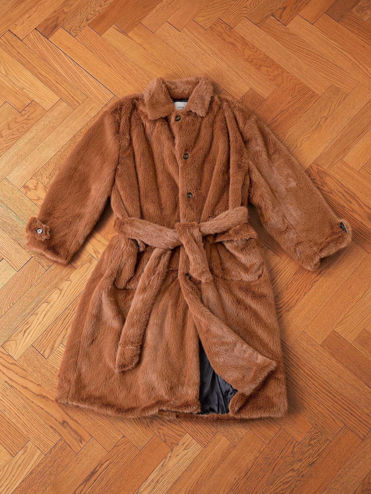 The Long Faux Fur Coat from FOUND is elegantly laid out on a wooden floor, showcasing its oversized fit with a belt and buttons.