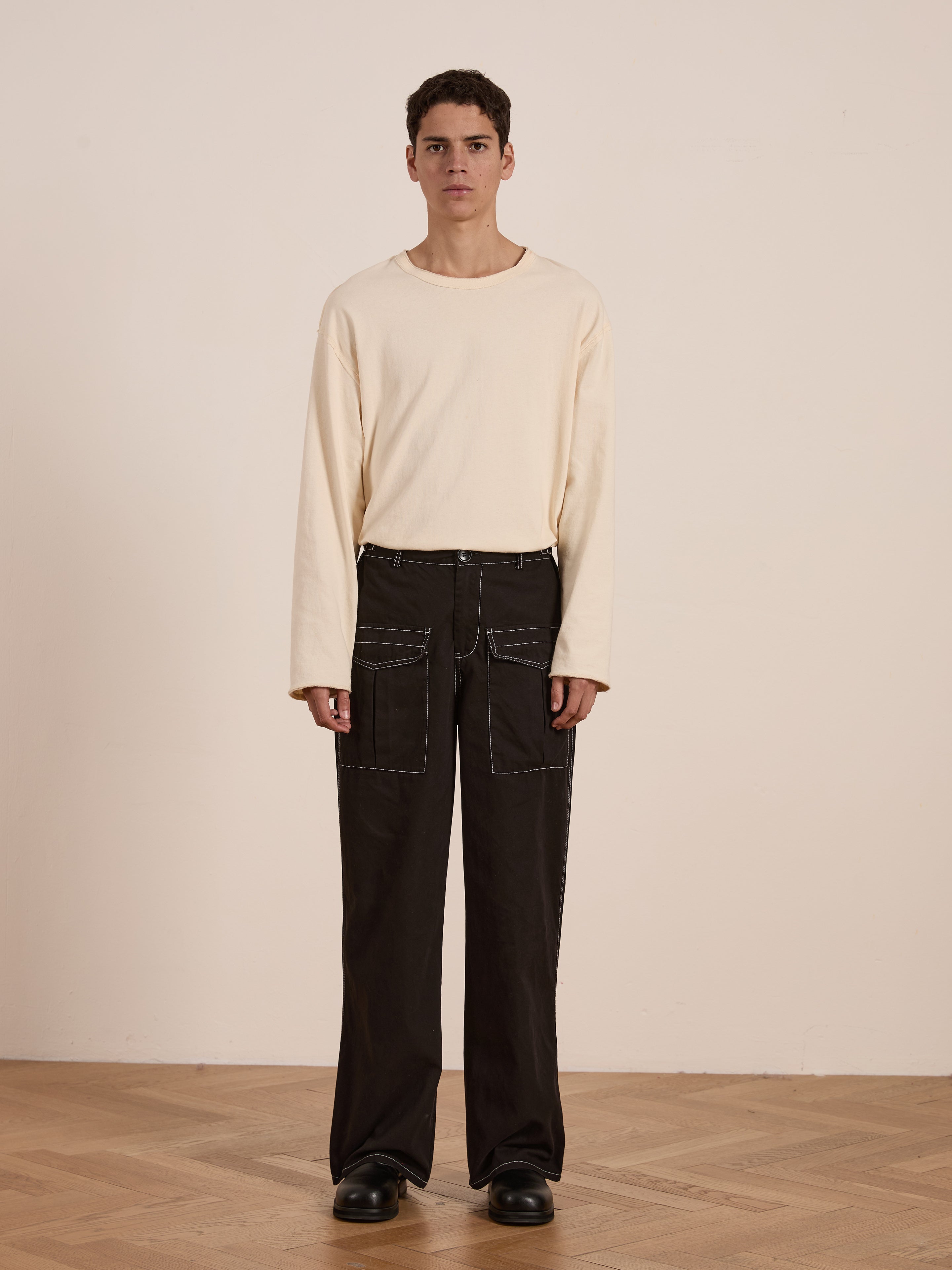 Indoors, an individual stands on a wooden floor dressed in a cream long-sleeve shirt and Black Lucca Contrast Pocket Pants by FOUND, featuring visible stitching, contrast pockets, and a relaxed fit; they are paired with black shoes.