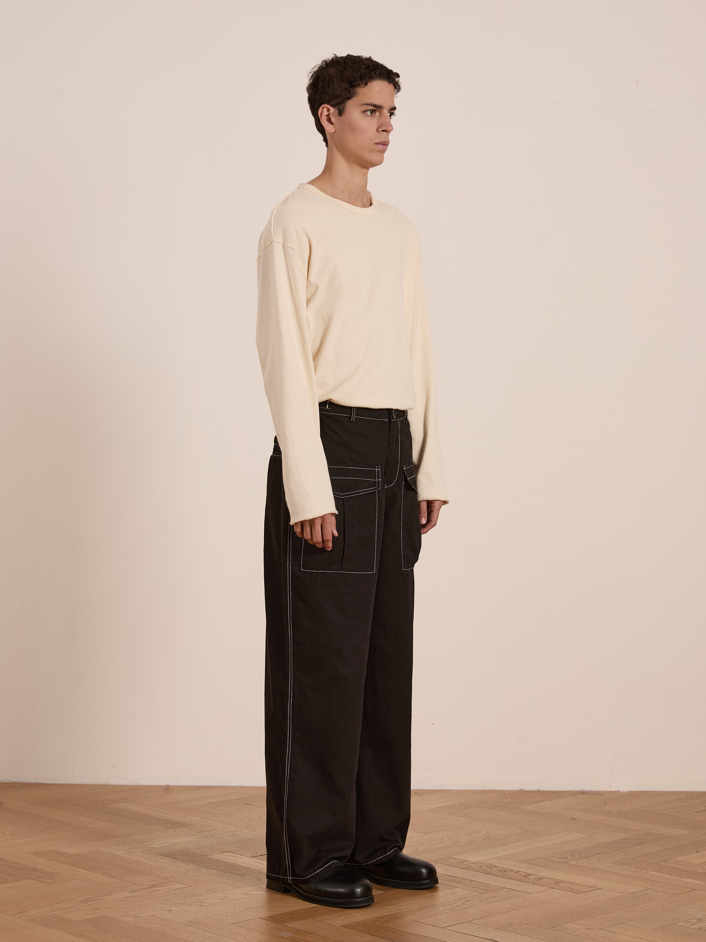 A person wearing a beige long-sleeve shirt and FOUND's Lucca Black Contrast Pocket Pants stands on a wooden floor against a plain wall.