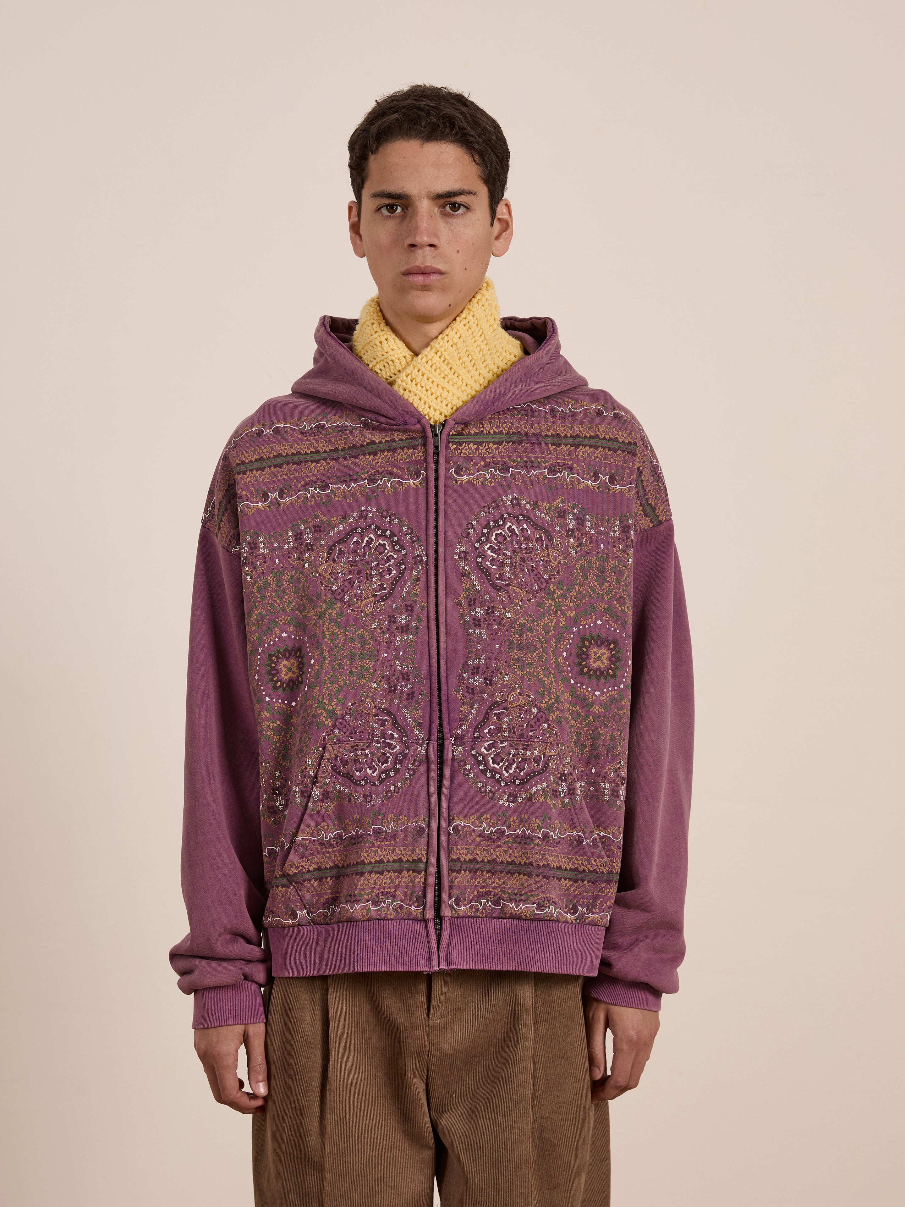 Wearing a streetwear staple, the person confidently sports a FOUND Motif Zip-Up Hoodie in purple pattern with a yellow knitted scarf against a plain background.