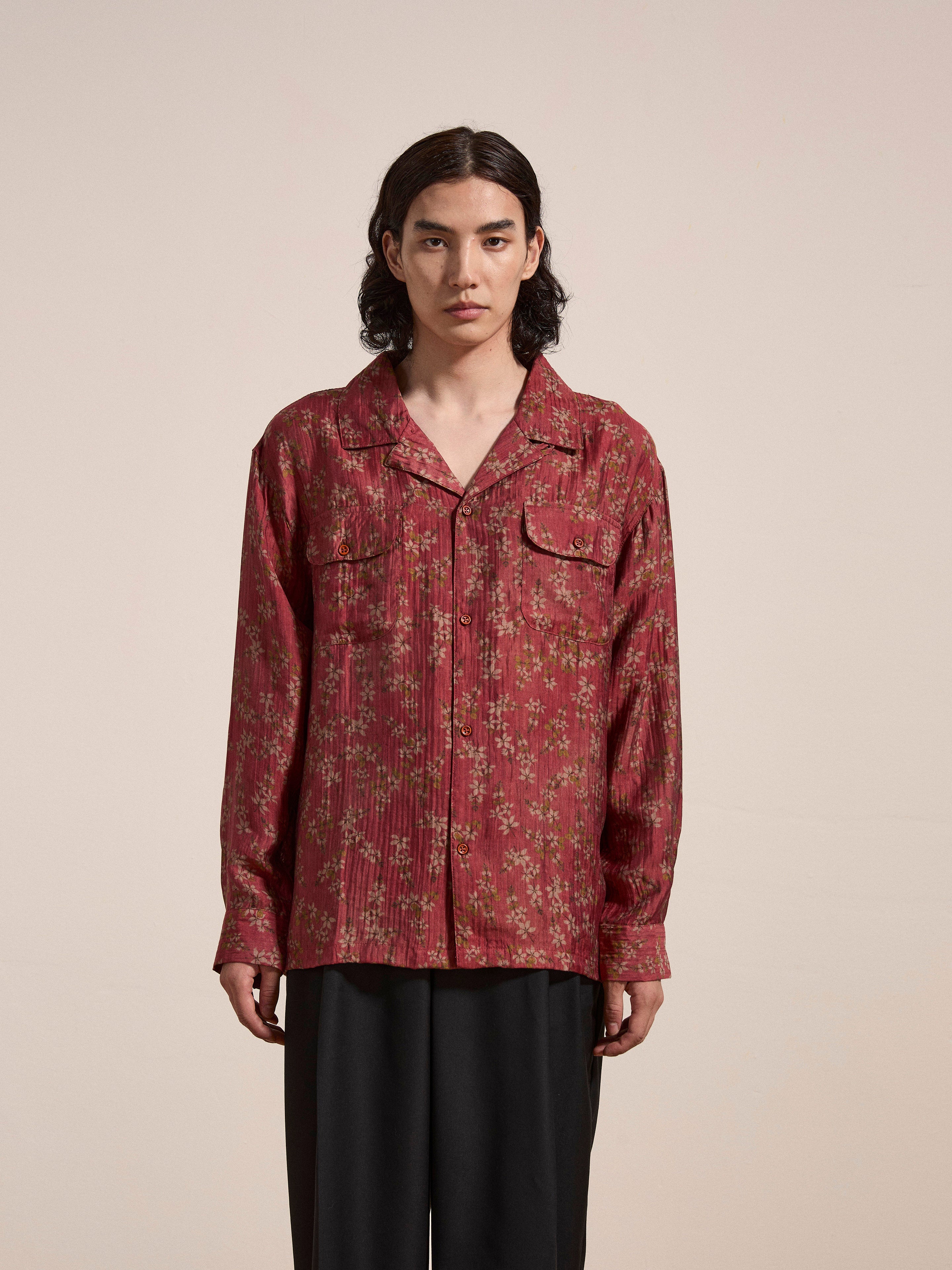 A person with shoulder-length dark hair wearing a red floral Charlot LS Camp Shirt by FOUND, featuring dual chest pockets, paired with black pants, standing against a plain background.