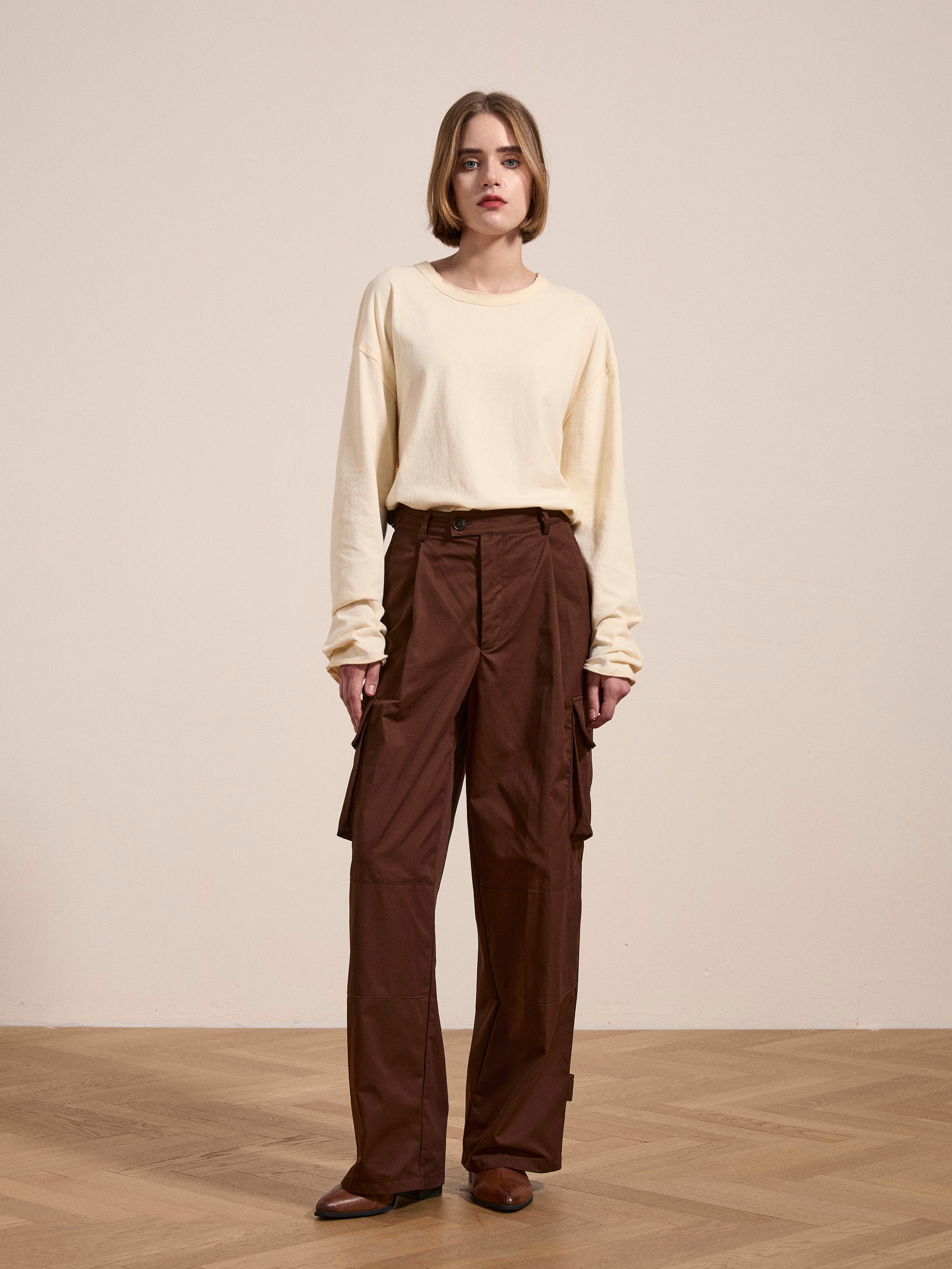 A person stands facing forward, wearing a beige long-sleeved shirt and FOUND's pleated pocket trousers in brown. The background is plain, featuring a light-colored wall and parquet flooring.