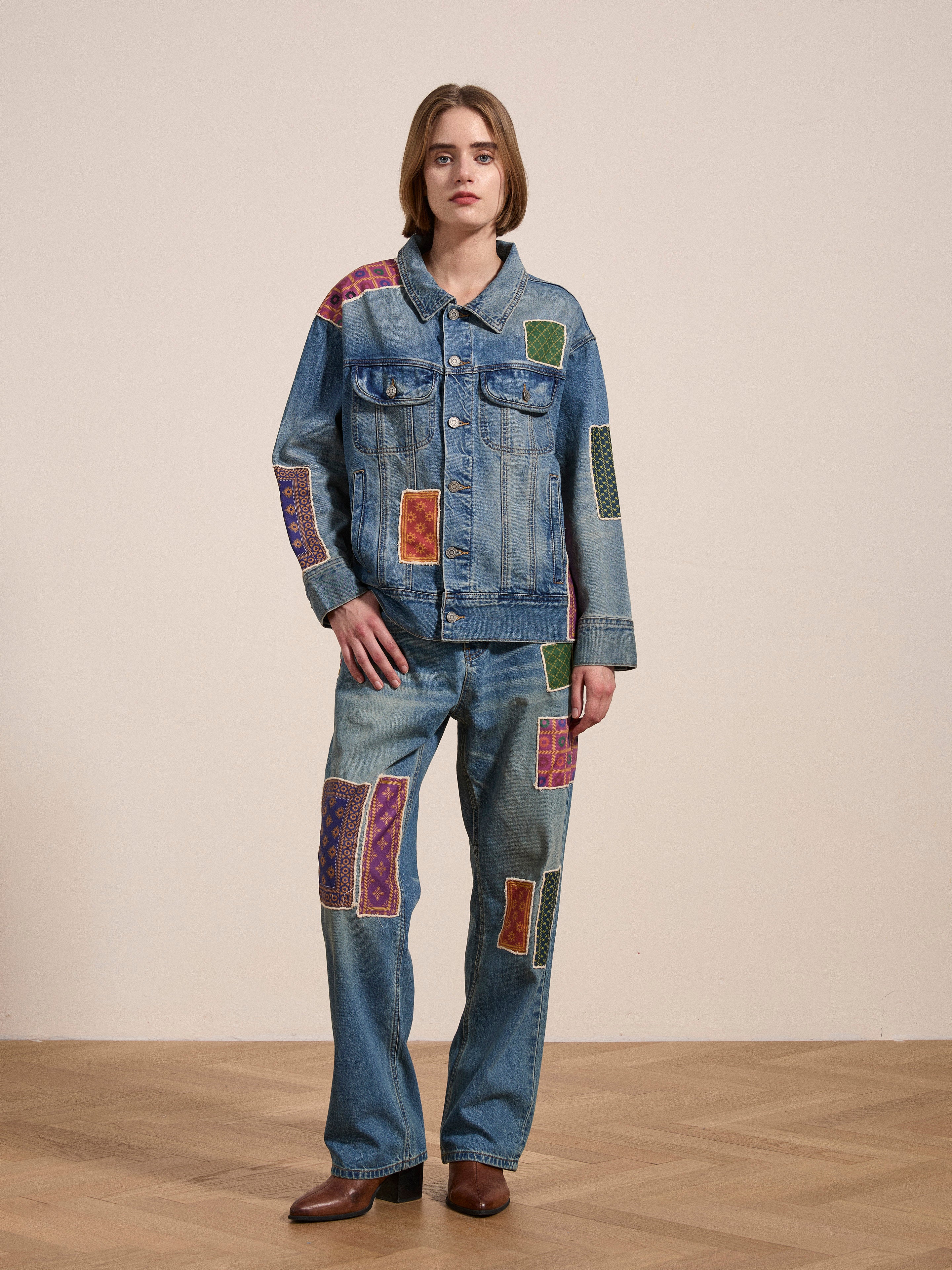 A person standing on a wooden floor, sporting a denim jacket and Found's Kantha Patchwork Jeans adorned with vintage blue wash and South Asian inspired patches, paired with brown shoes. The background is plain and light-colored.