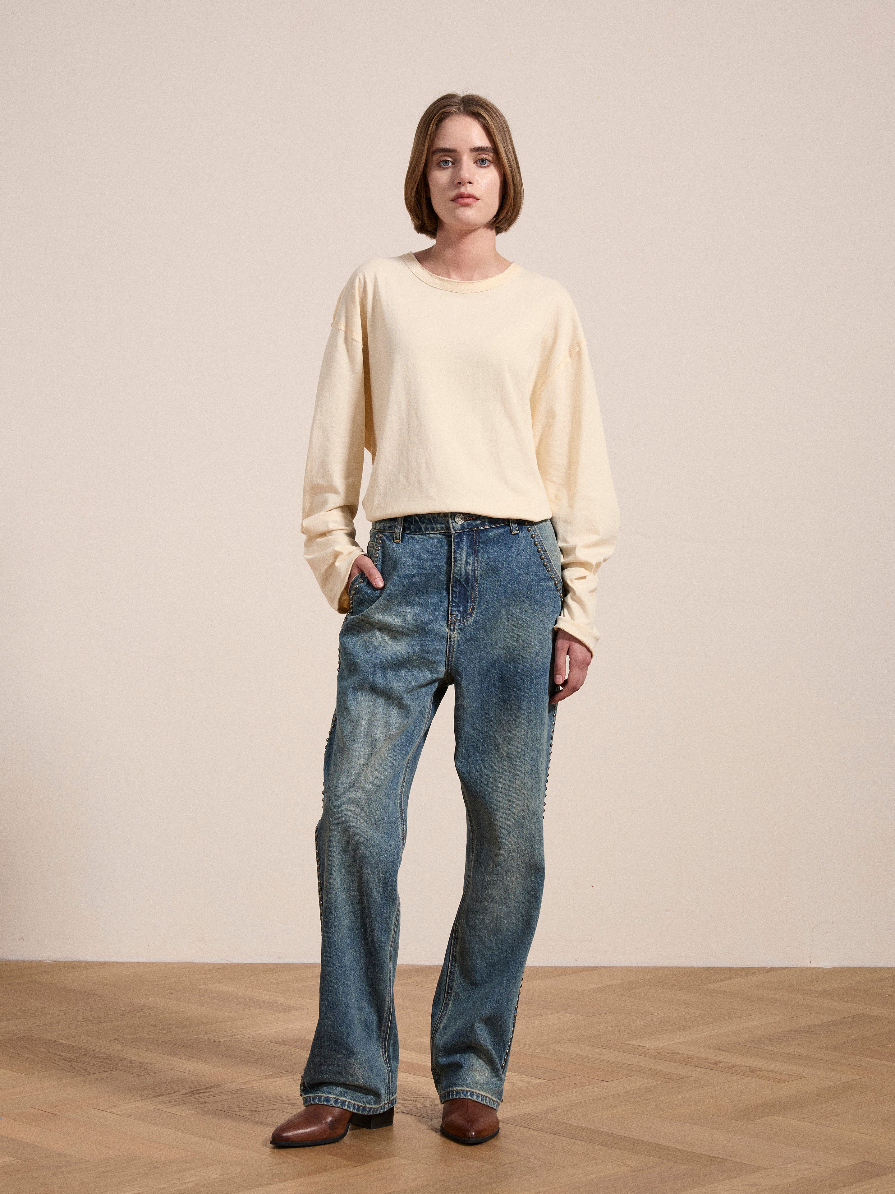 A person stands on a wooden floor wearing a lightweight, long-sleeved shirt, FOUND Azar Studded Jeans adorned with antique matte silver studs, and brown shoes, with one hand in their pocket.