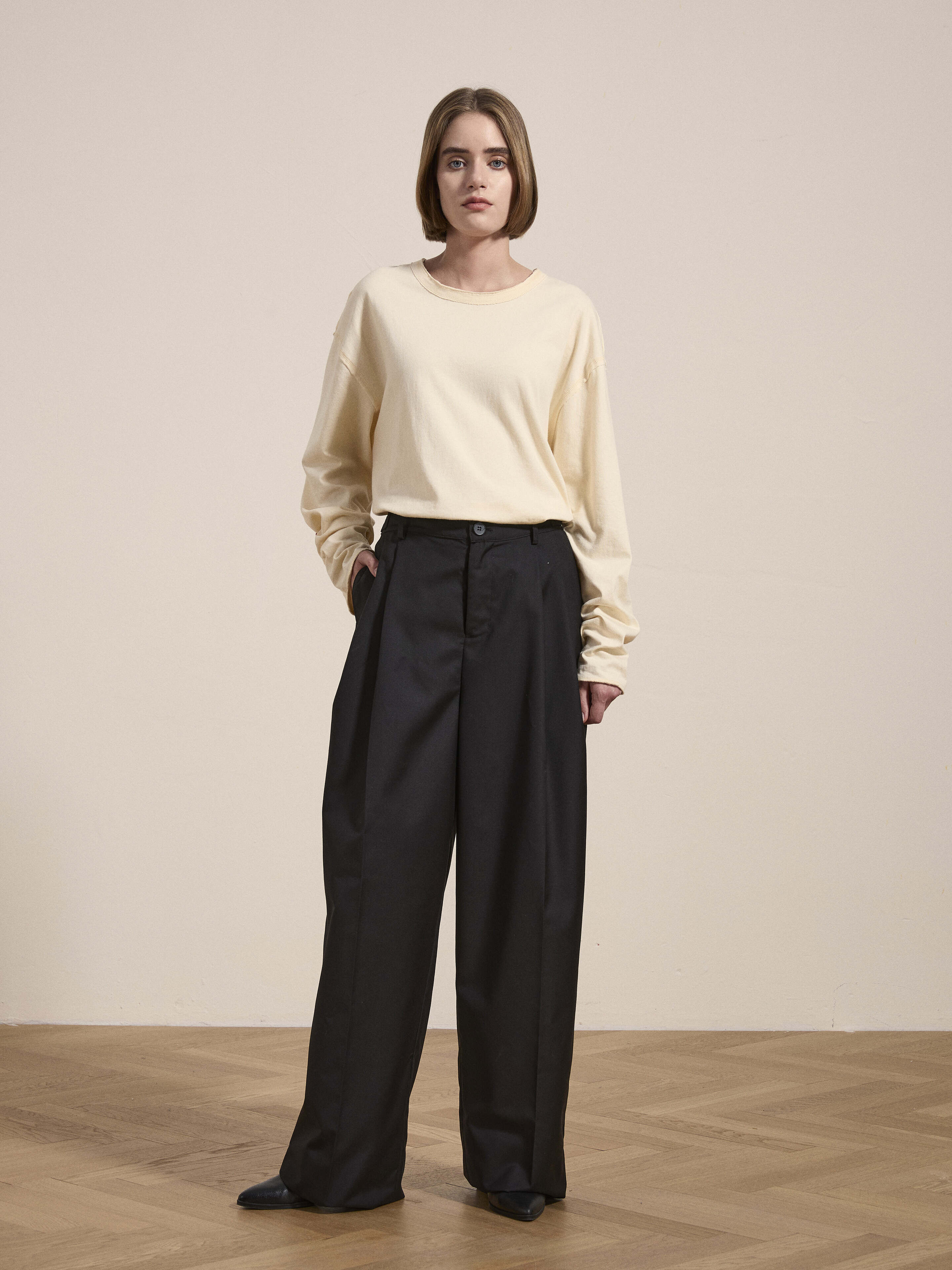 A person with a bob haircut stands on a wooden floor, wearing a cream-colored long-sleeve shirt and high-waisted, wide-leg Pleated Trousers by Found.