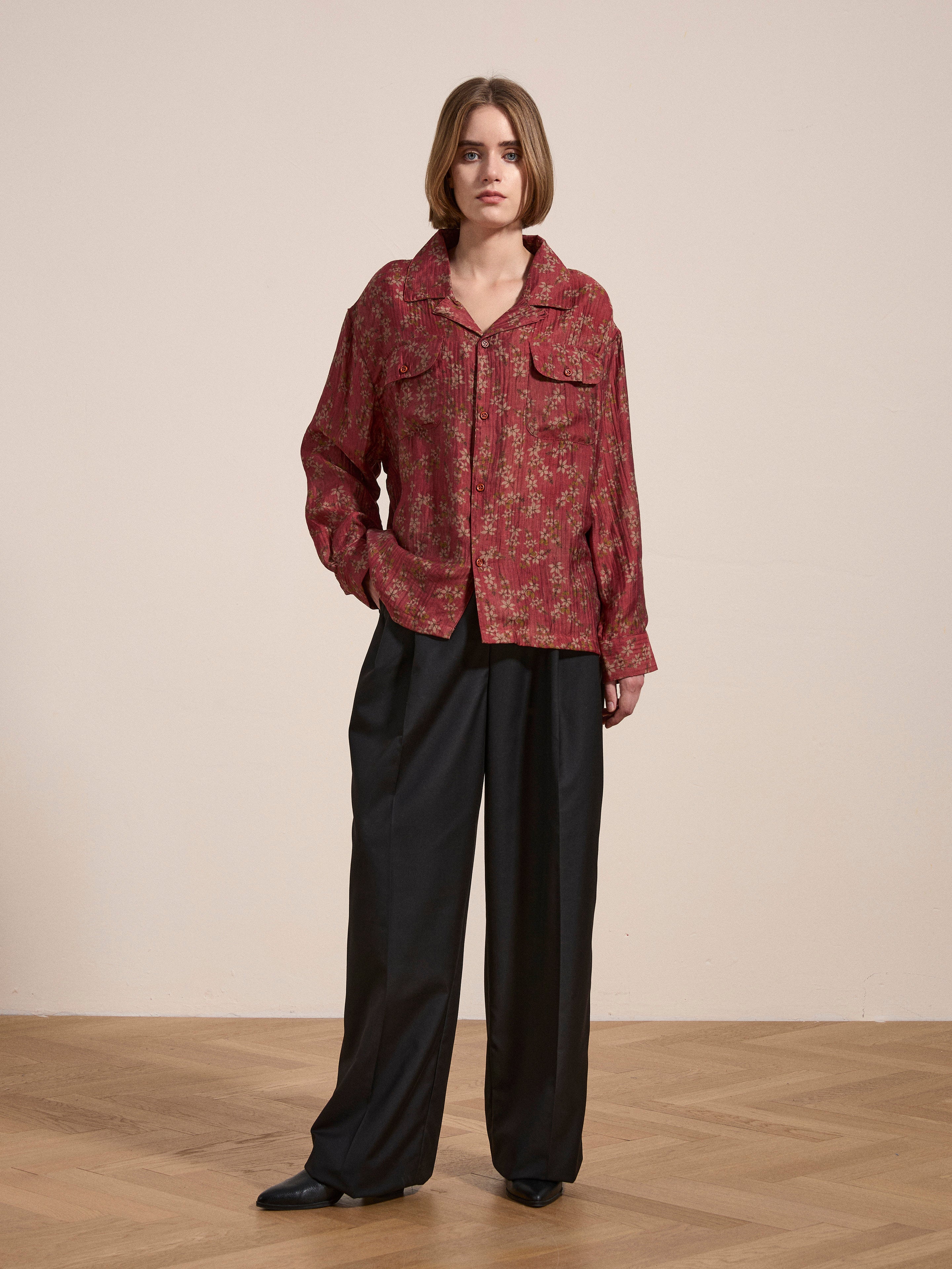 A person stands indoors on a wooden floor, wearing the FOUND Charlot LS Camp Shirt featuring red floral patterns and dual chest pockets, paired with black wide-leg trousers.