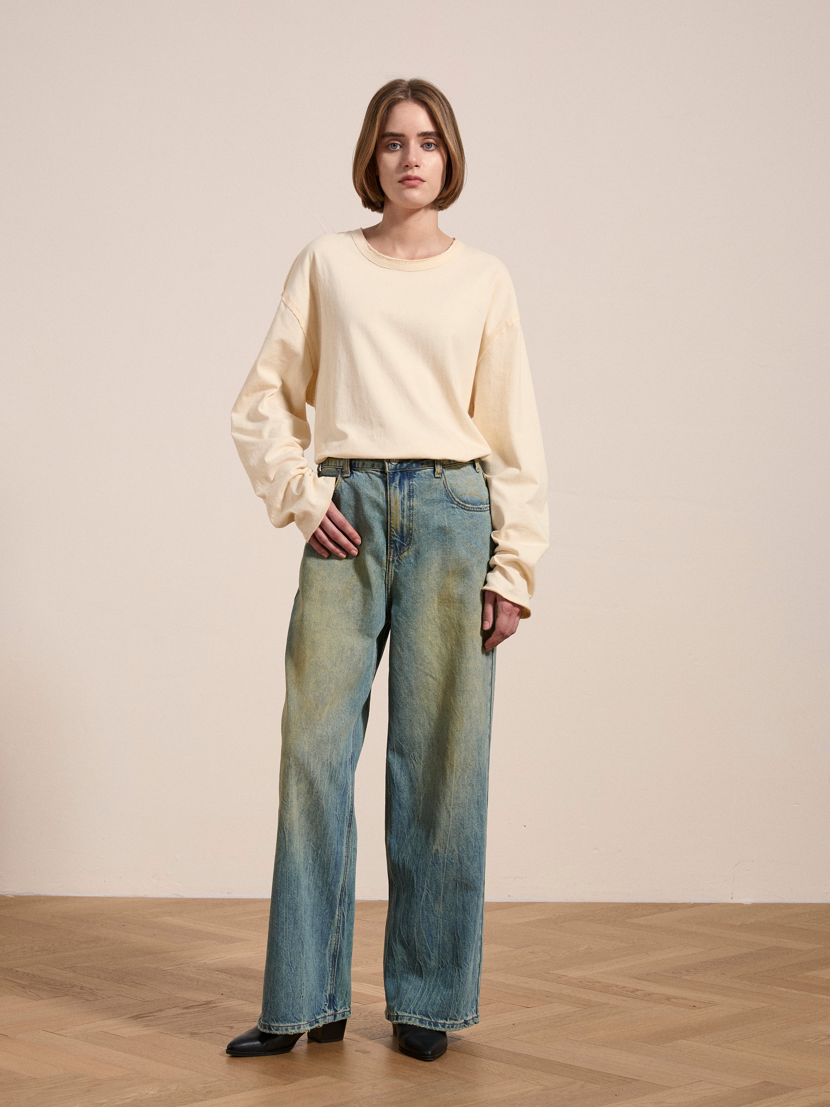 A person with short hair stands on a wooden floor, wearing a beige long-sleeved shirt and the Lacy Baggy Jeans by FOUND, which feature a loose fit and light distressing.