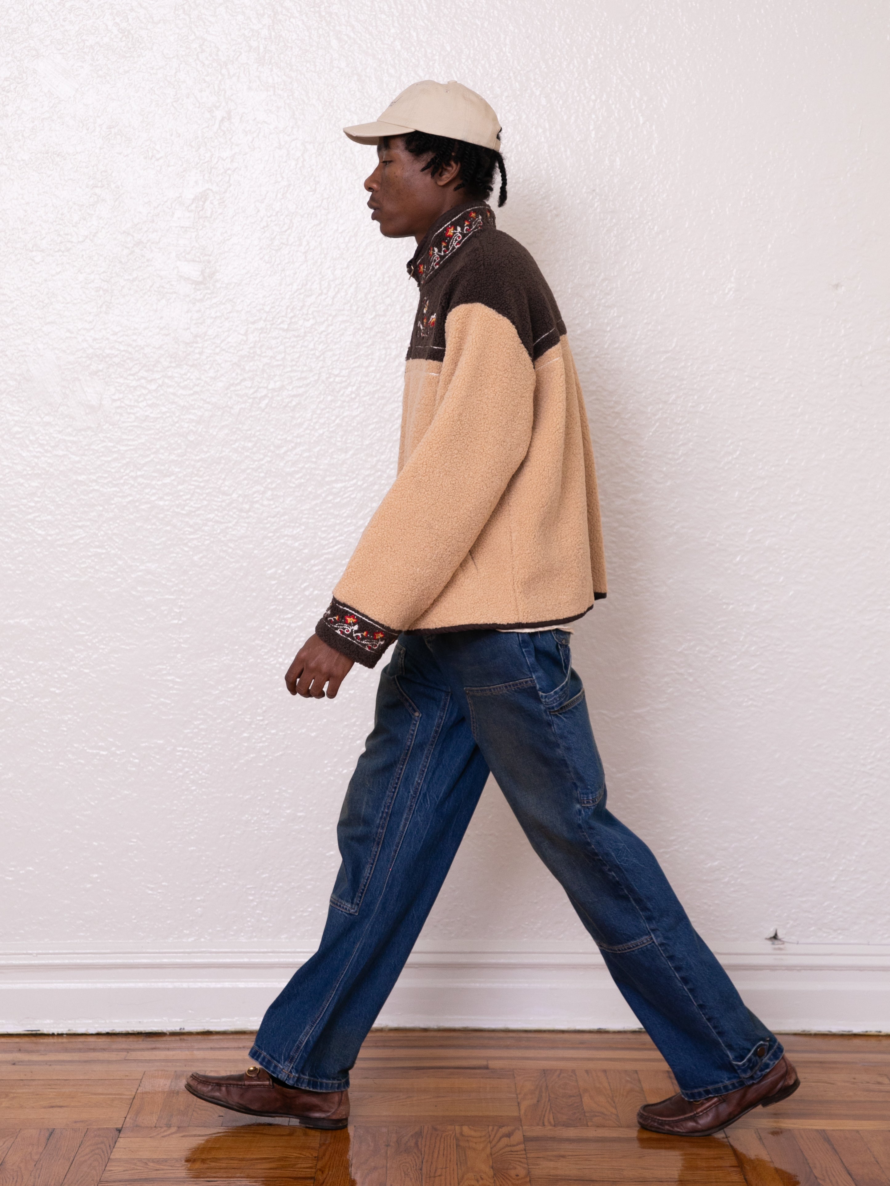 A person wearing a Pahari Floral Embroidered Sherpa Pullover by FOUND, blue jeans, and a cream cap with delicate Pahari embroidery walks against a plain white wall.