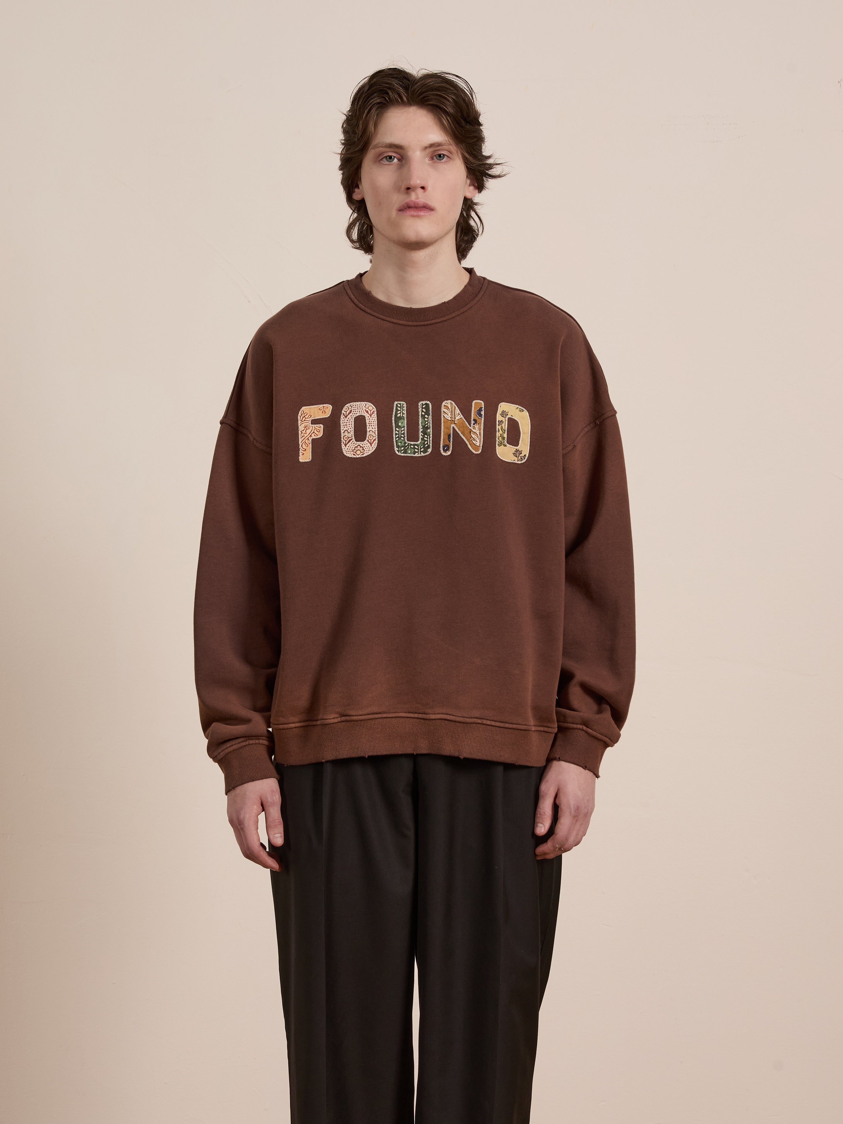 A person with medium-length hair wears the FOUND Patterned Logo Crewneck sweatshirt in brown against a neutral background.