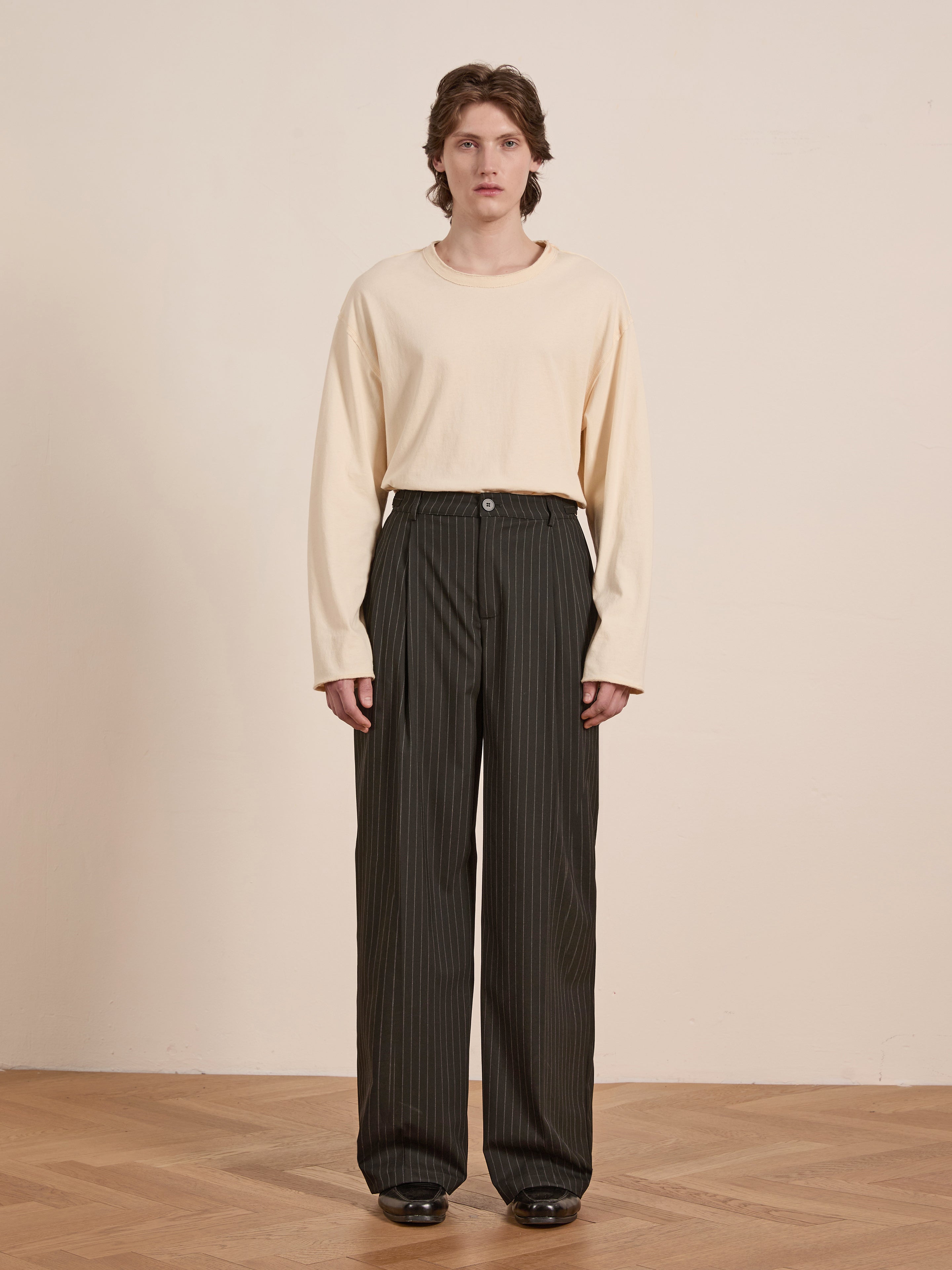 Against a plain background, a person wears a light long-sleeve shirt paired with FOUND's Pinstripe Pleated Trousers—dark, relaxed fit, high-rise with vertical stripes—standing on a wooden floor.