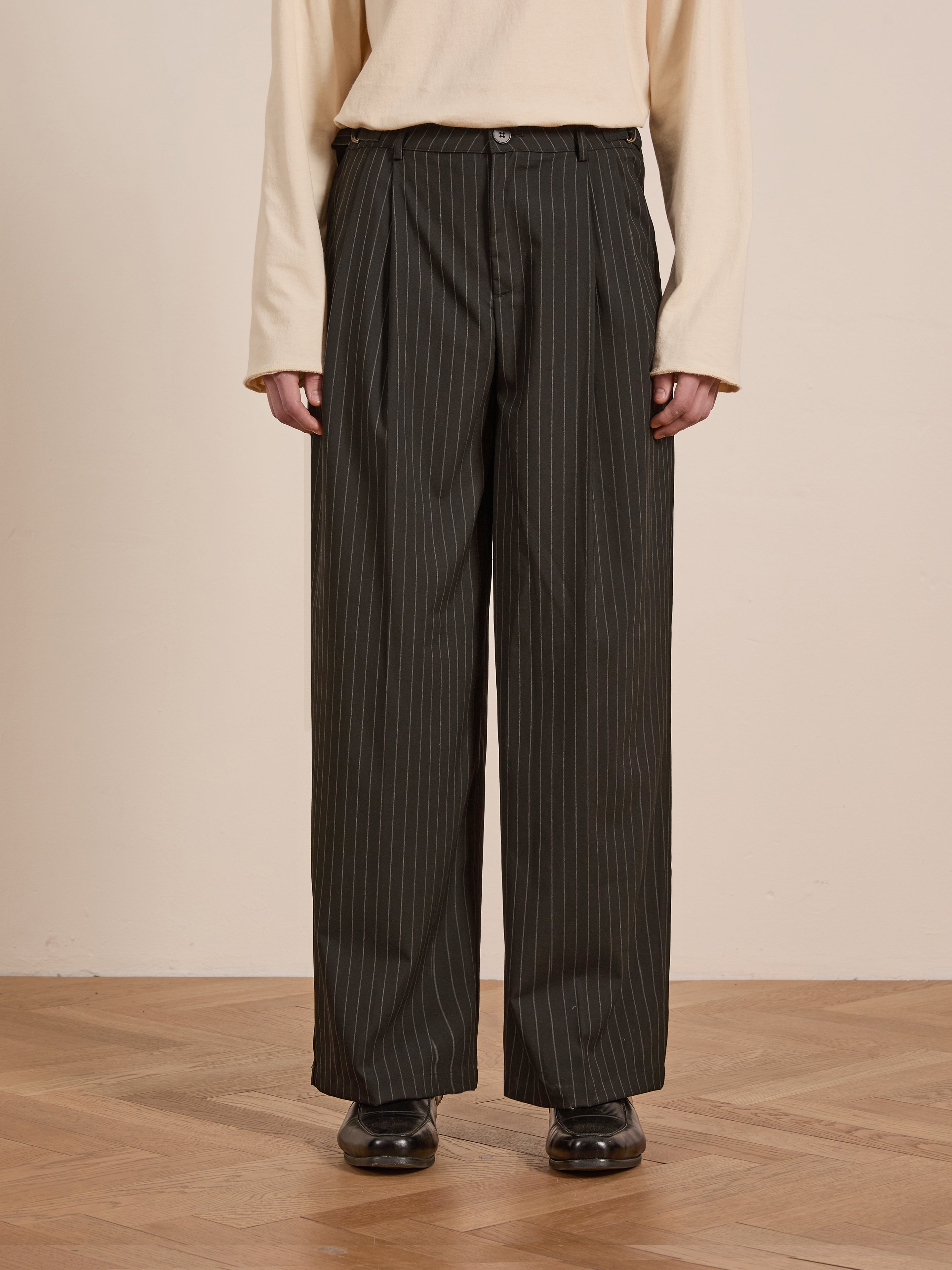 A person in a beige long-sleeve shirt and FOUND's high-rise Pinstripe Pleated Trousers with a relaxed fit stands on a wooden floor.