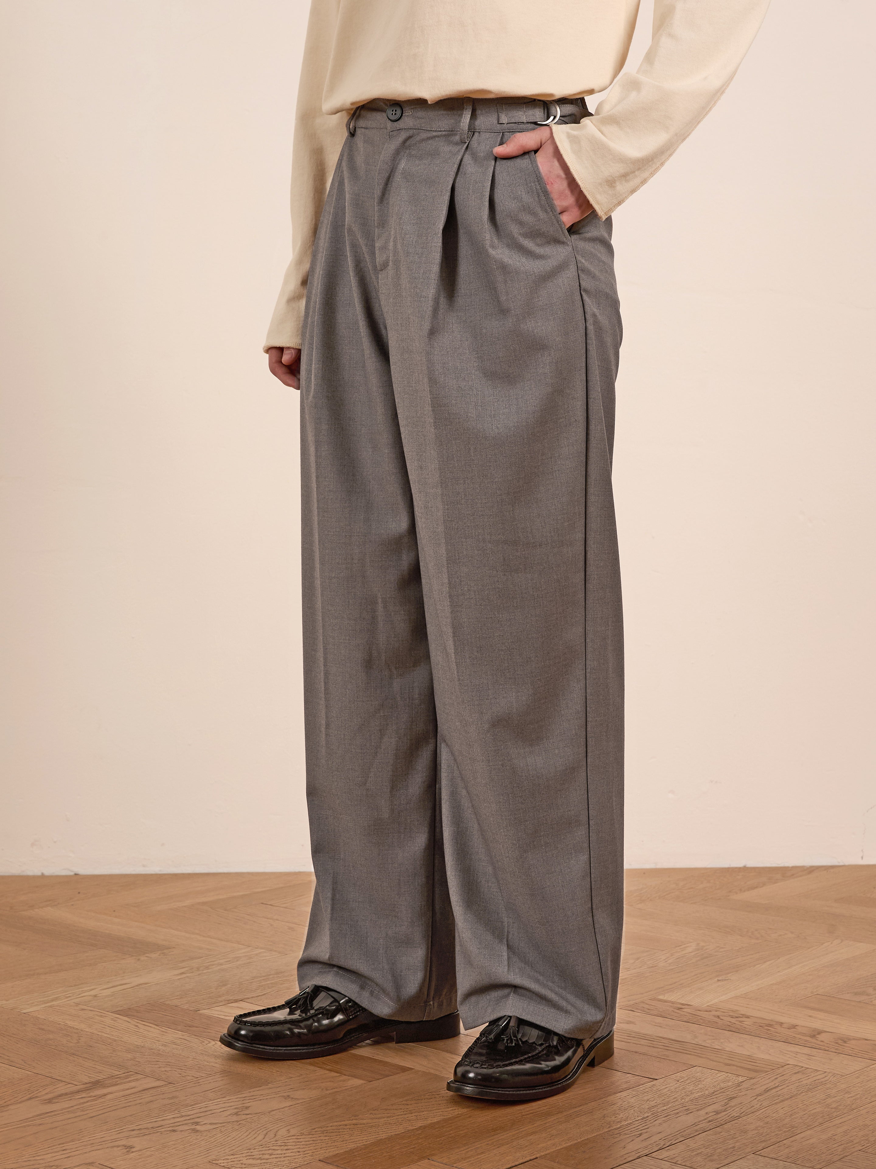 A person in a cream shirt and FOUND Pleated Trousers in gray, featuring a high-rise design and adjustable waist tabs, stands on a wooden floor, paired with sleek black shiny shoes.