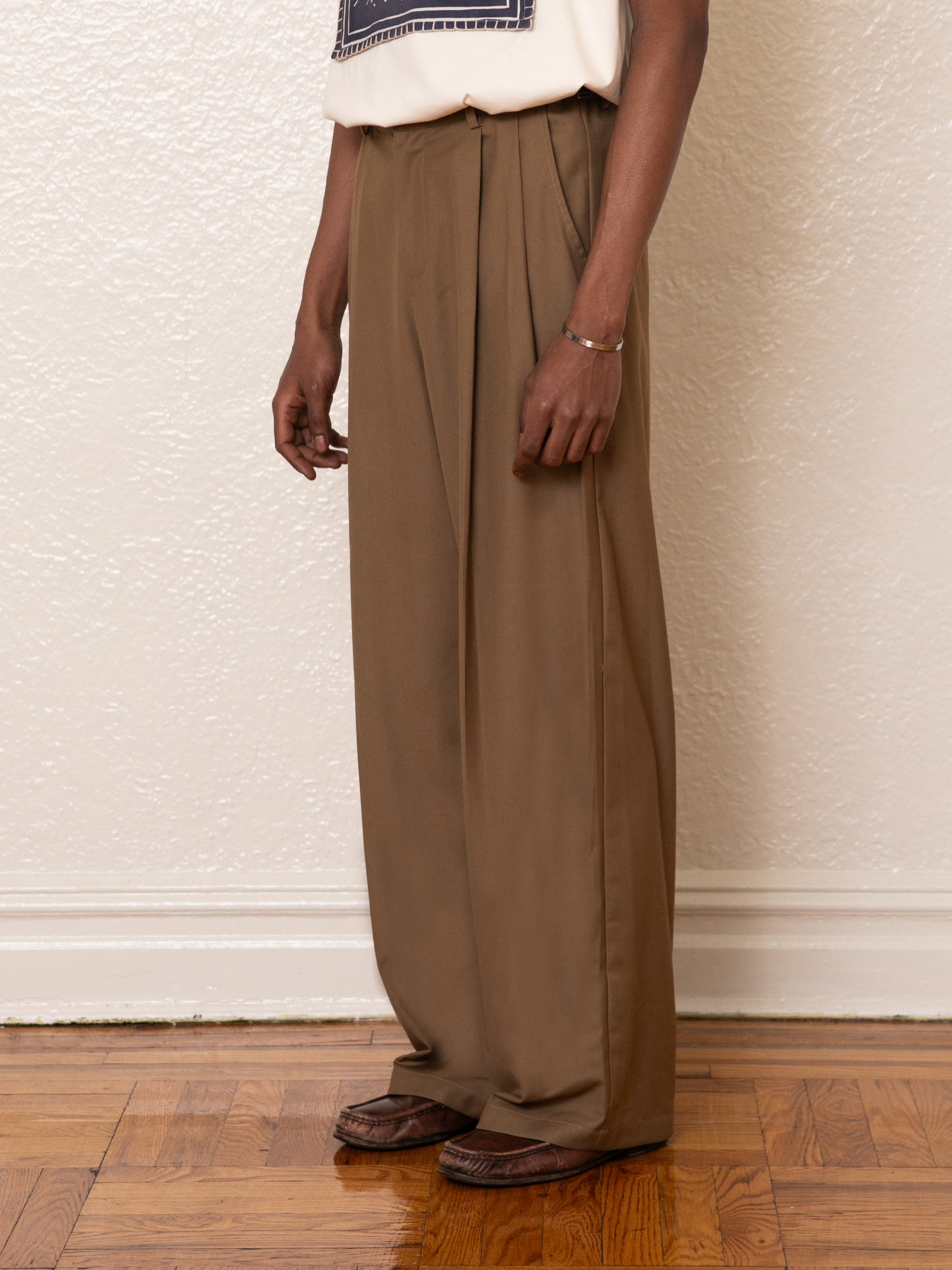 A person in a cream shirt and FOUND Pleated Trousers with an adjustable waist stands before a textured beige wall on a wooden floor.