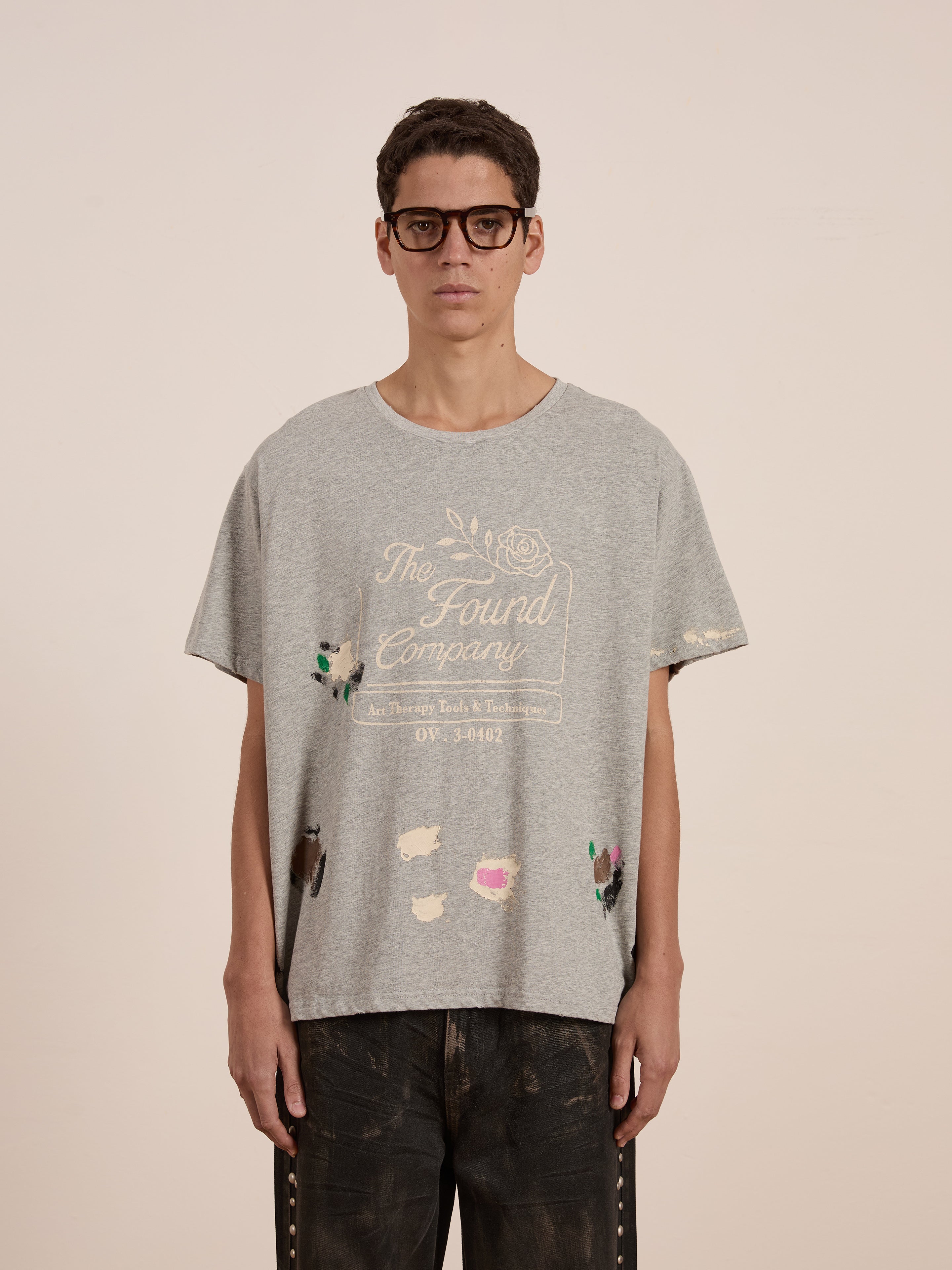 Person in glasses and dark pants wearing FOUND's Painters Company Tee in grey, featuring a vintage-inspired graphic with paint splatters.