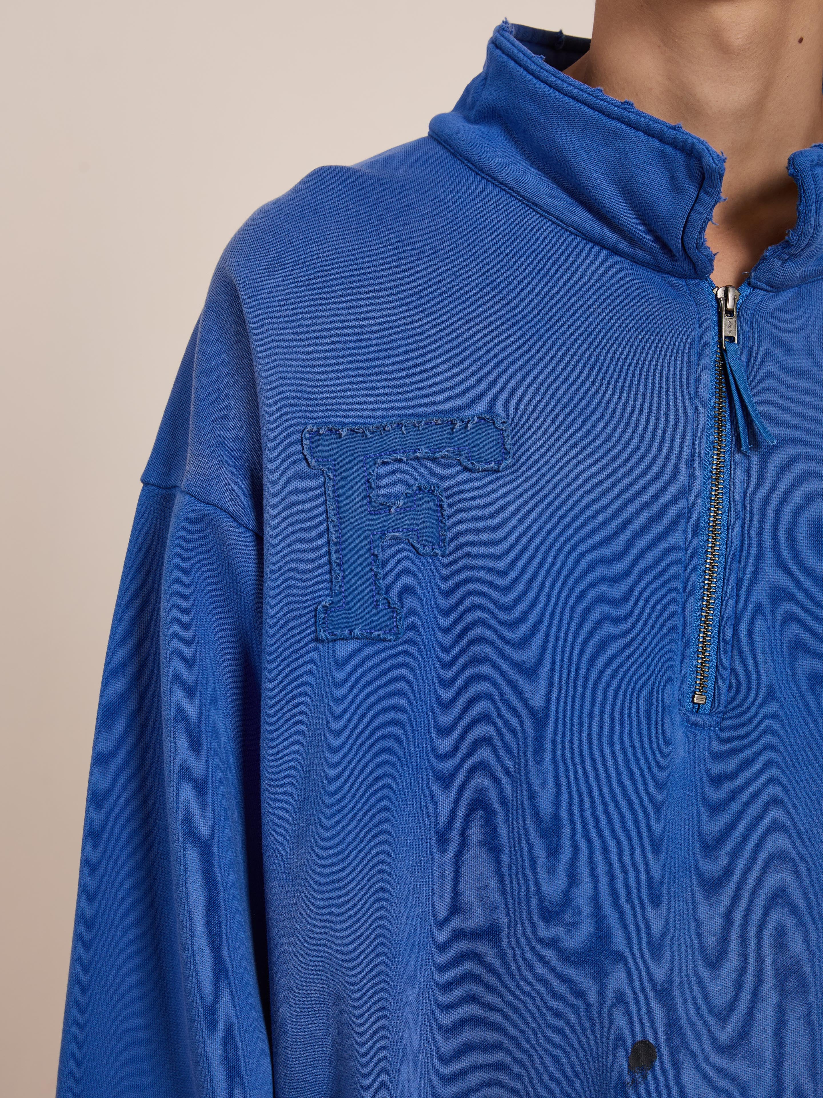 A person is dressed in FOUND's Painters Pullover Sweatshirt, a blue unisex design featuring a frayed "F" chest patch and partial collar zipper, combining casual comfort with a workwear-inspired style.