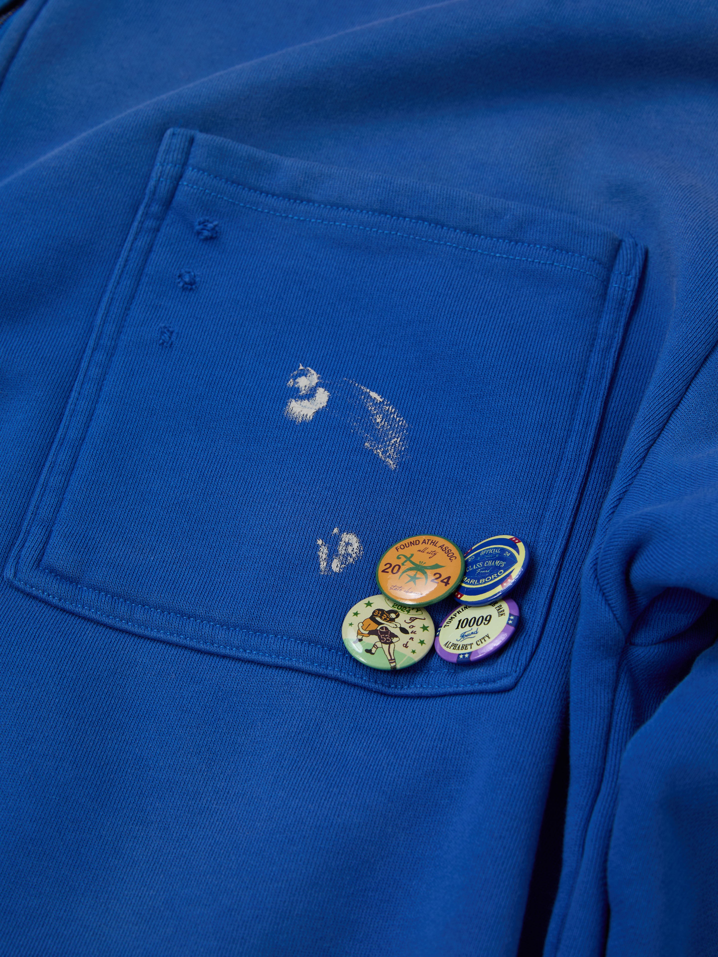 FOUND's Painters Pullover Sweatshirt is a unisex blue design with a faded pocket patch and four vibrant pins, adding a unique twist to classic workwear fashion.