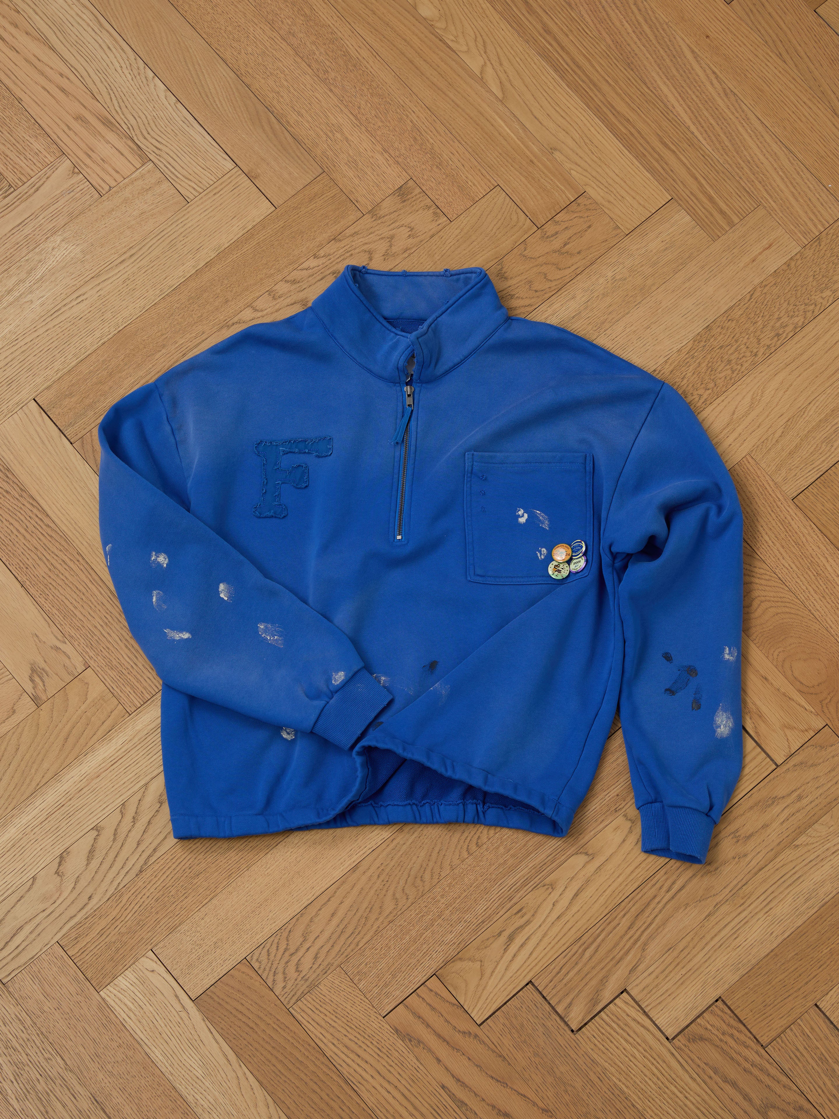 The FOUND Painters Pullover Sweatshirt is a blue, unisex workwear piece with a quarter-zip, chest pocket adorned with badges, sleeve paint-like marks, and an "F" design on the front. It rests on a wooden floor.