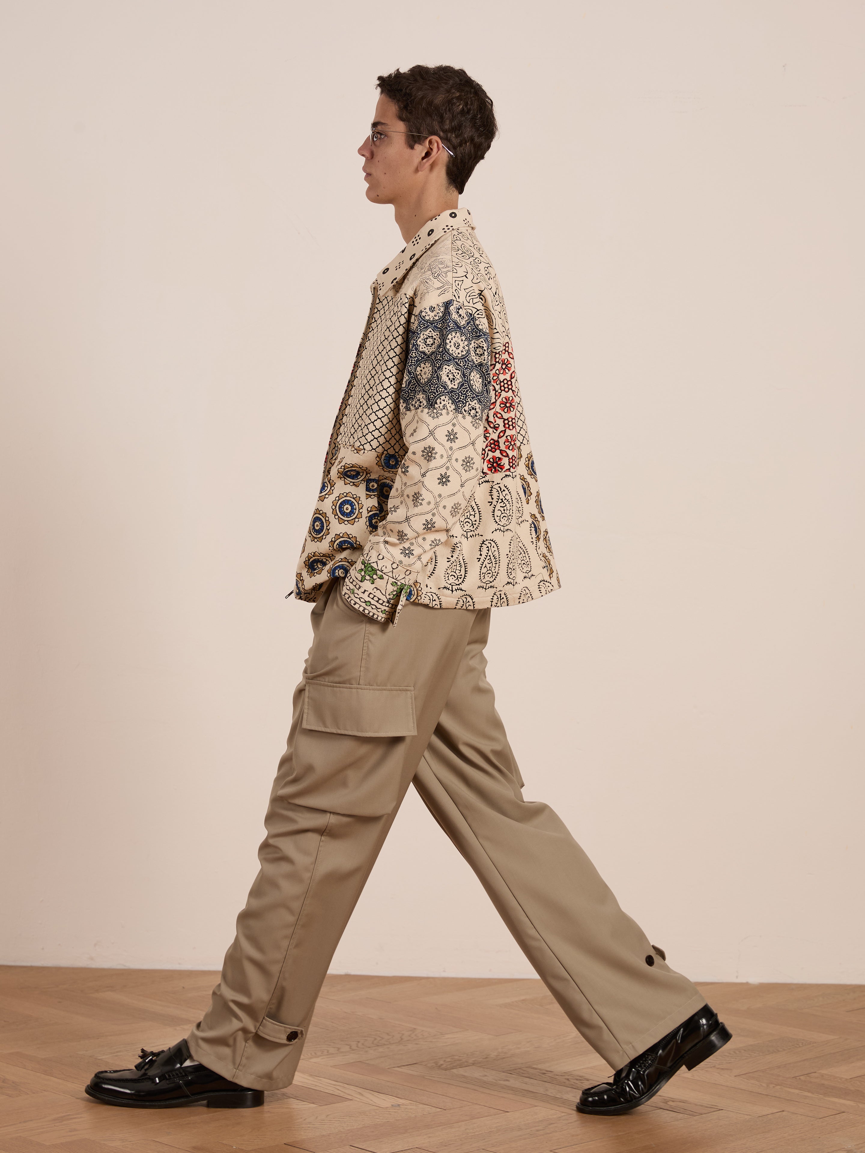 A person strides sideways on a wooden floor, wearing the FOUND Paisley Mosaic Work Jacket with beige cargo pants and sleek black shoes. The unisex outfit adds effortless style to their step.