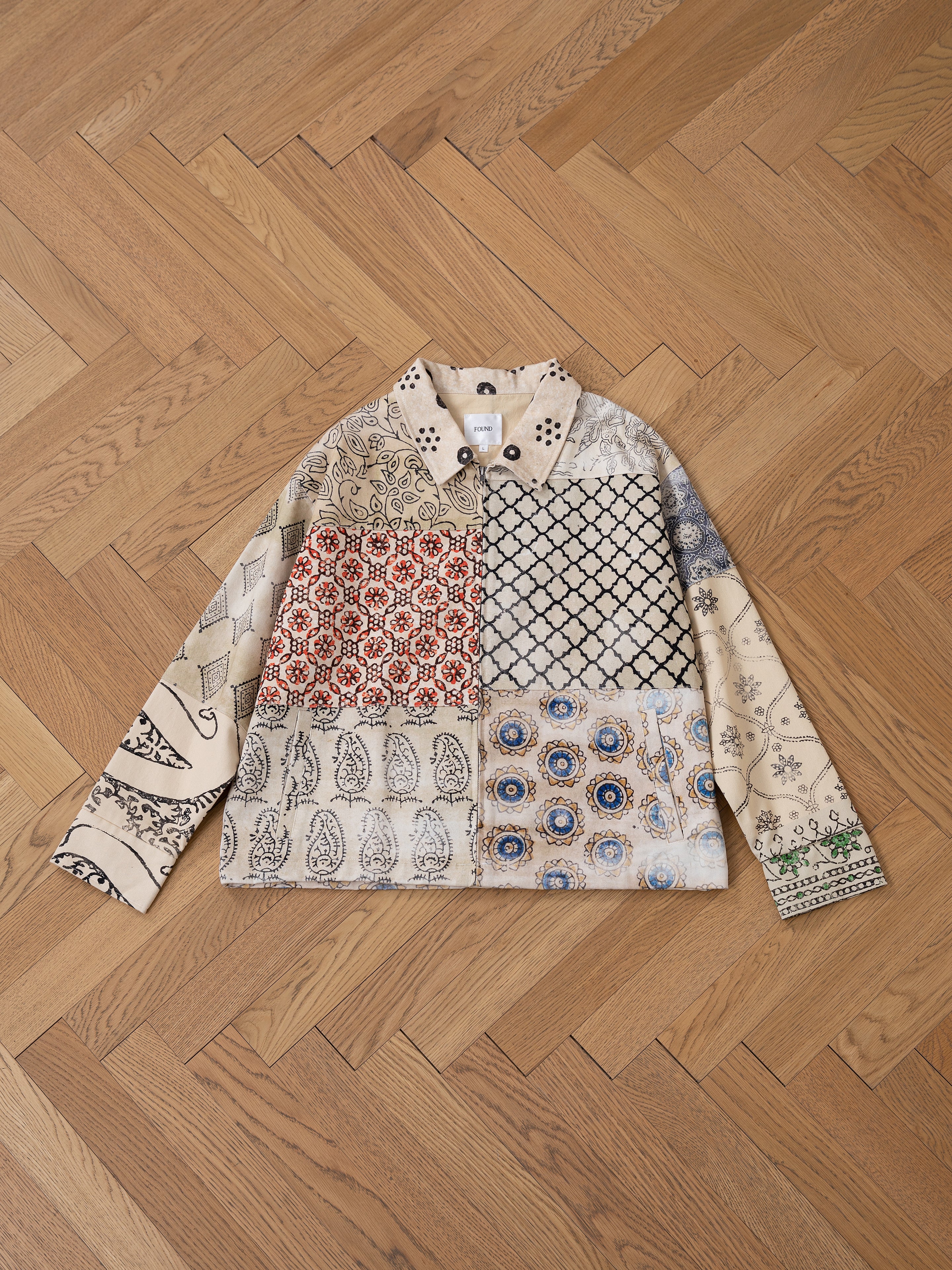 The FOUND Paisley Mosaic Work Jacket showcases a unisex design with intricate patterns and earthy colors, highlighted by hints of paisley mosaic, displayed on a chevron-patterned wooden floor.