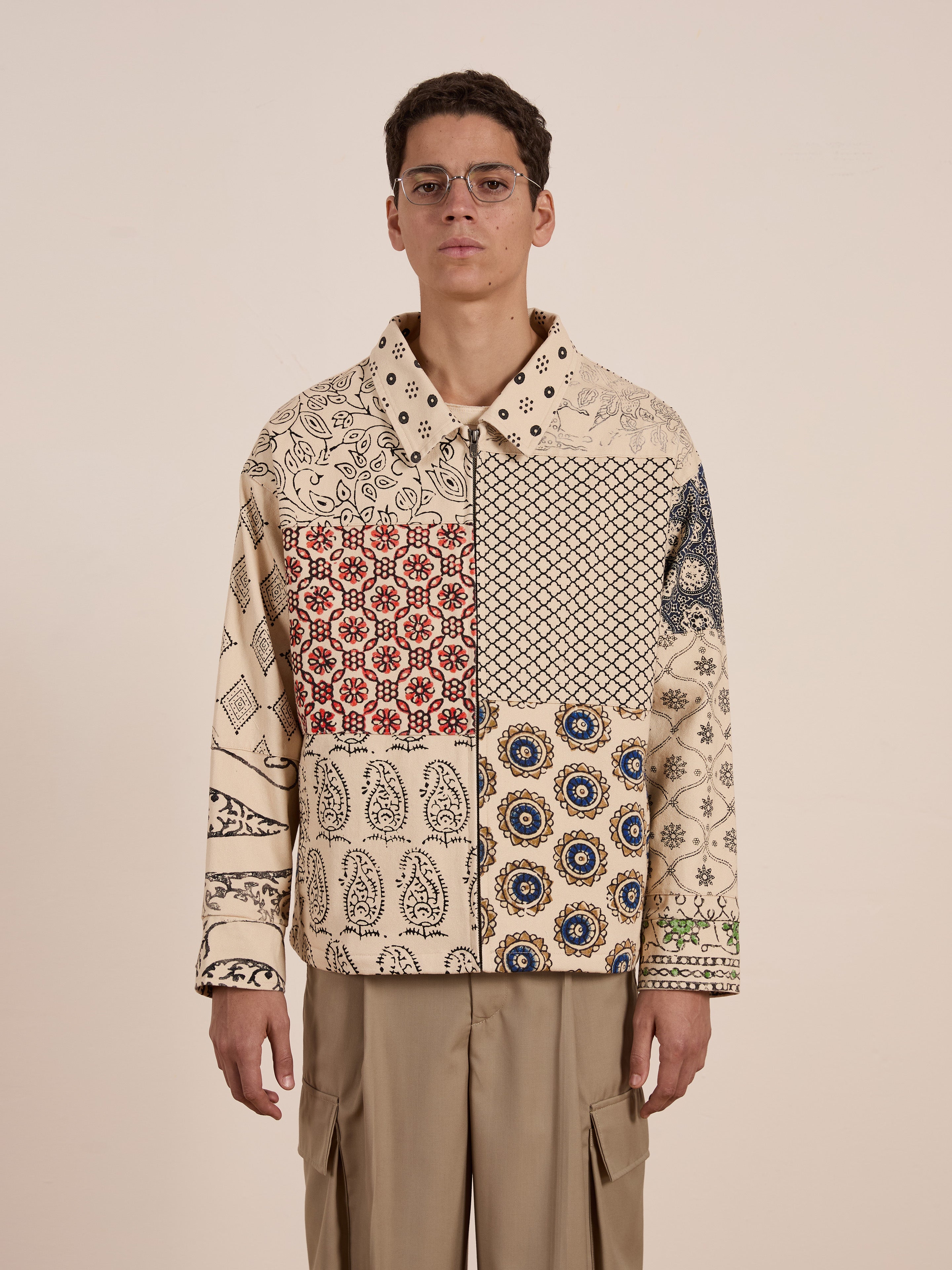 A person in glasses sports the unisex Paisley Mosaic Work Jacket by FOUND, showcasing a beige, red, and blue patchwork pattern with tan trousers against a simple backdrop.