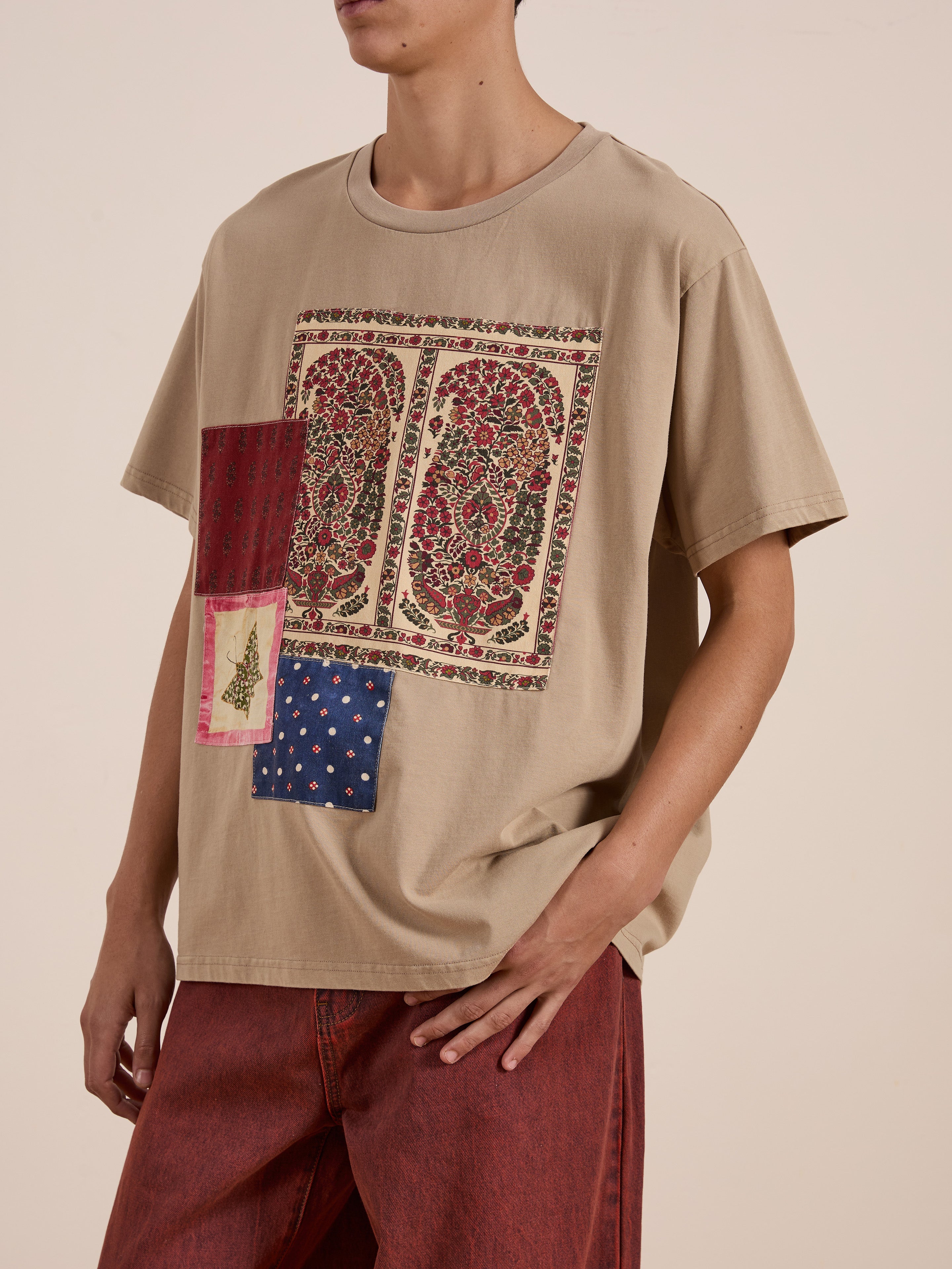 A person stands against a neutral background wearing FOUND's Patchwork Tee in beige, featuring intricate Kantha embroidery in red and blue, paired with vibrant red pants.