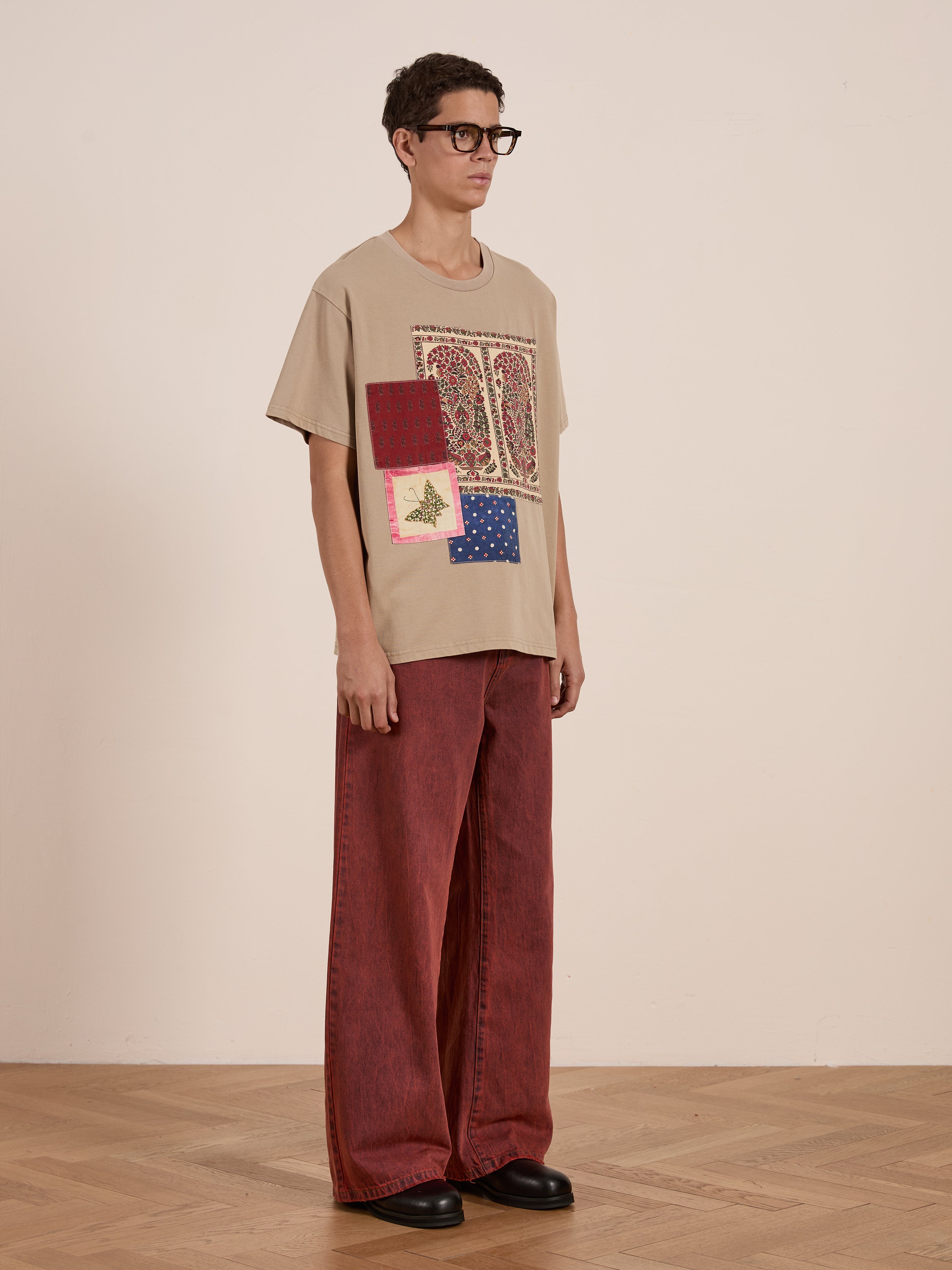 A person wearing the FOUND Patchwork Tee, a beige graphic T-shirt inspired by South Asian quilts, paired with red wide-leg pants and black shoes, stands elegantly on a wooden floor.