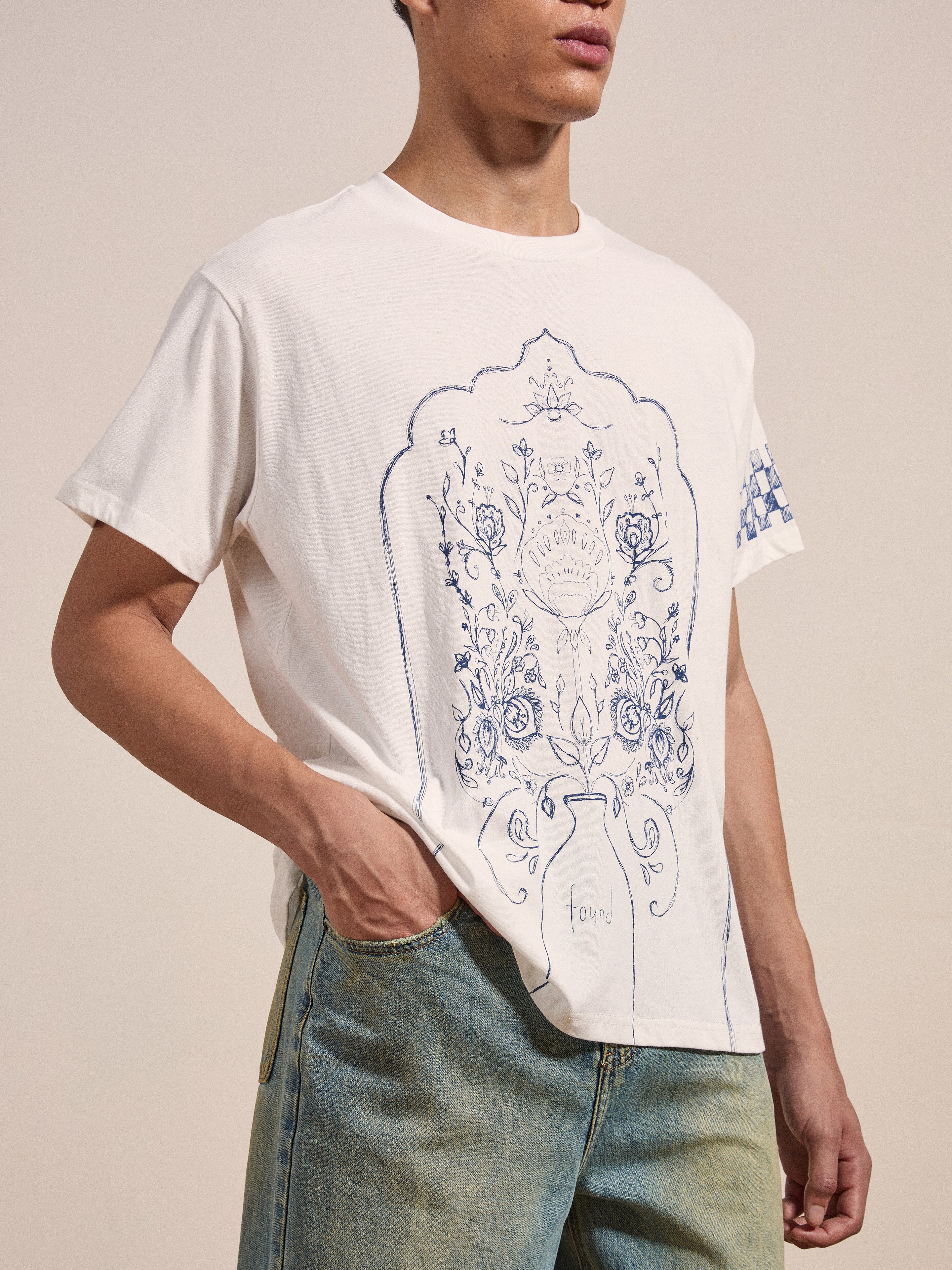 A person wearing the FOUND Pen Flower Medley T-shirt, featuring a detailed blue floral and geometric design on the front, paired with light blue jeans.