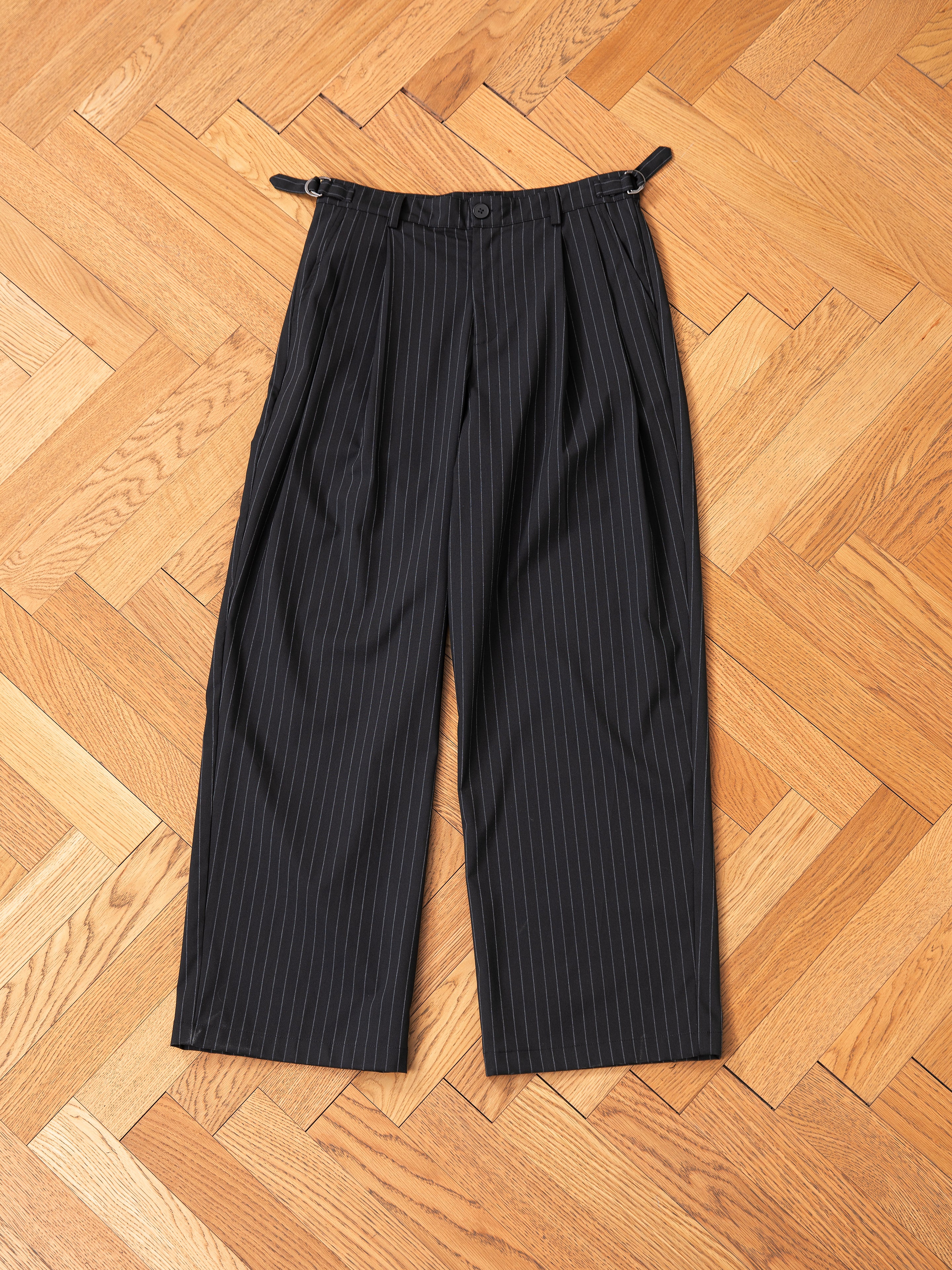 The Pinstripe Pleated Trousers by FOUND, featuring a wide-leg design with double pleats at the front and adjustable waist tabs, are laid flat on a herringbone wood floor.