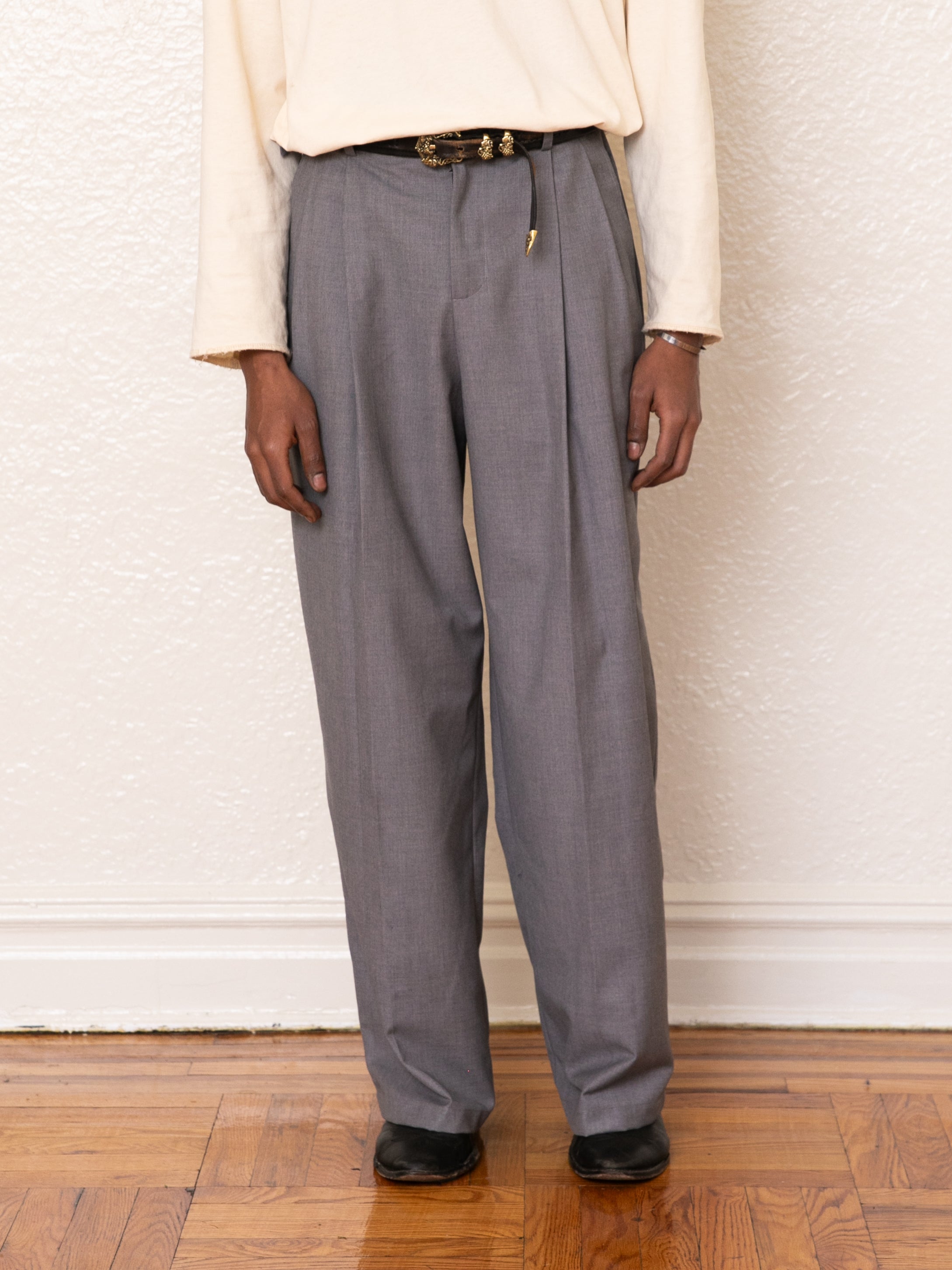 A person poses against a textured wall wearing stylish gray FOUND Pleated Trousers with a high-rise fit, a cream long-sleeve shirt, a black belt, and sleek black shoes.