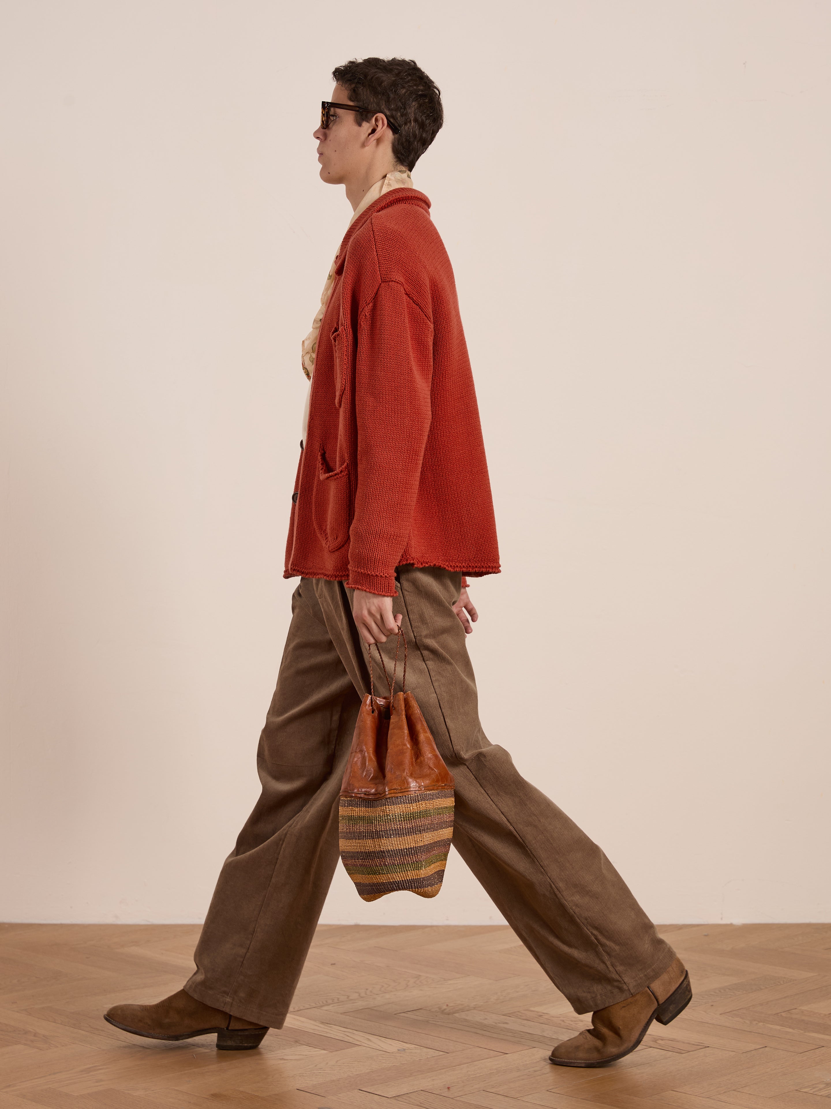 An individual wearing a Hunter Knit Collared Cardigan by FOUND in red and brown trousers moves elegantly with a striped bag, ideal for transitional layering.