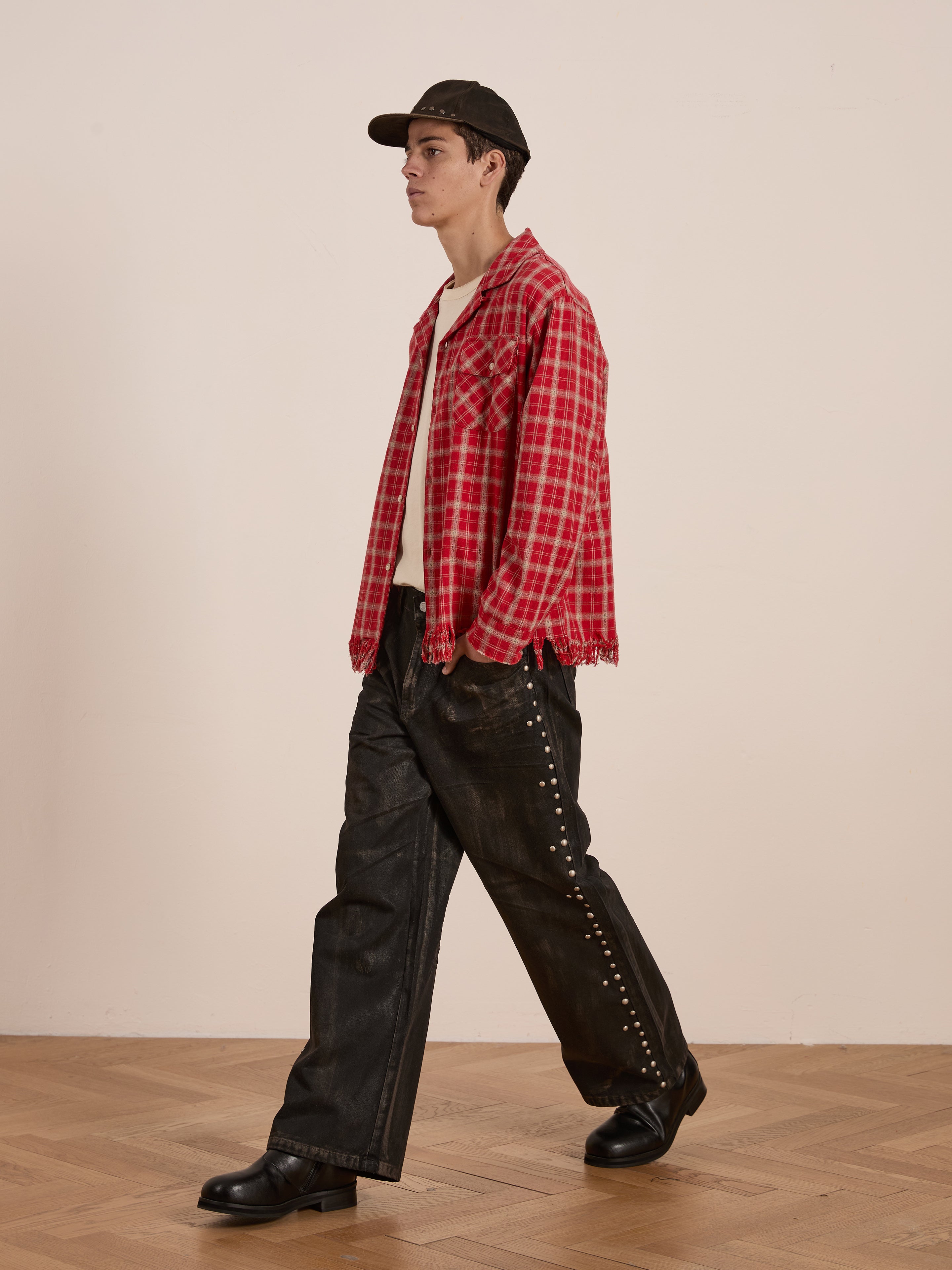 Someone in a Raw Cut Fringe Plaid Camp Shirt by FOUND, black pants, boots, and a black cap walks across a wooden floor against a plain backdrop.