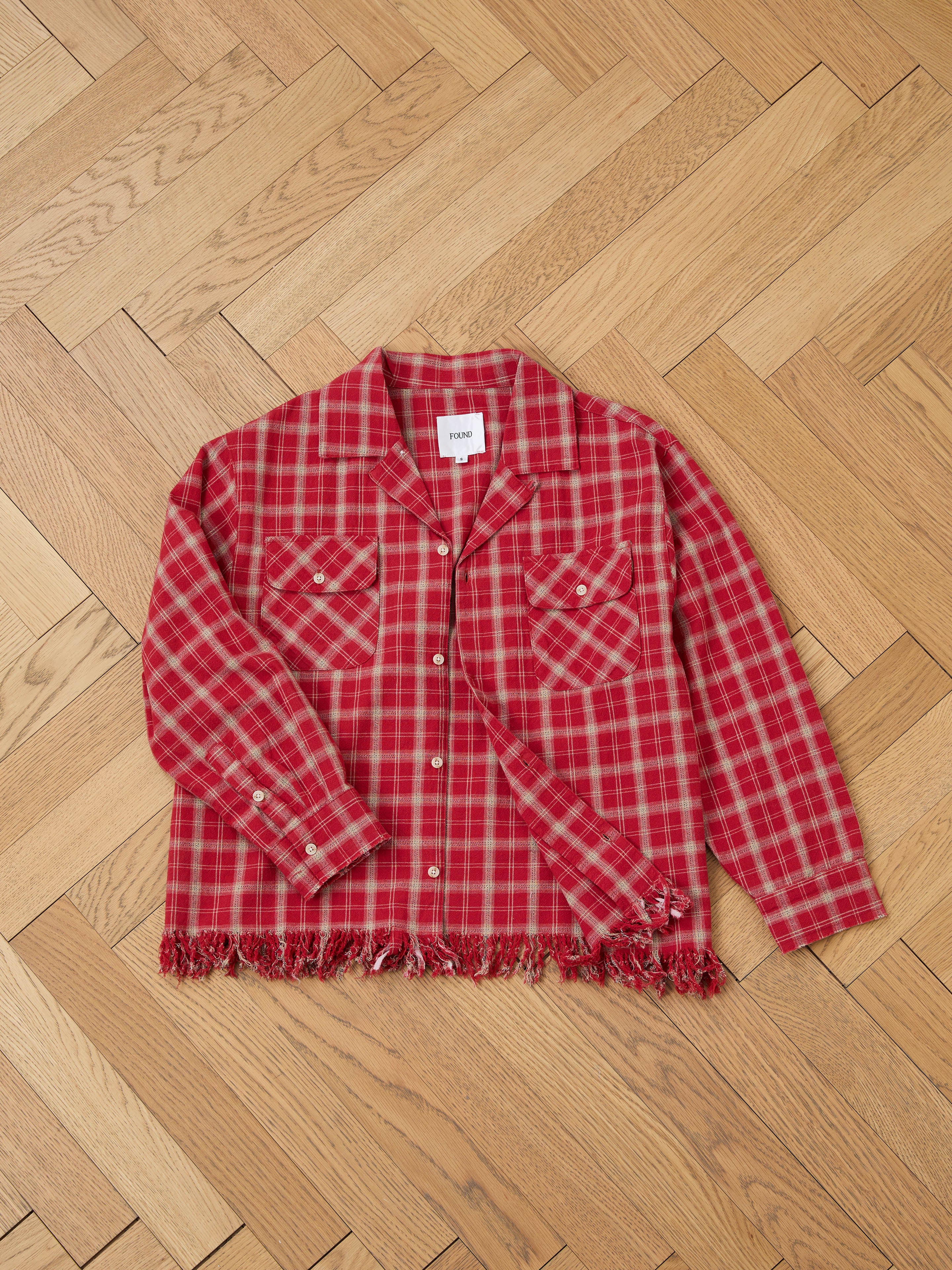 The FOUND Raw Cut Fringe Plaid Camp Shirt rests on a herringbone-patterned wooden floor, delivering the cozy comfort of flannel with its red hue and frayed edges.