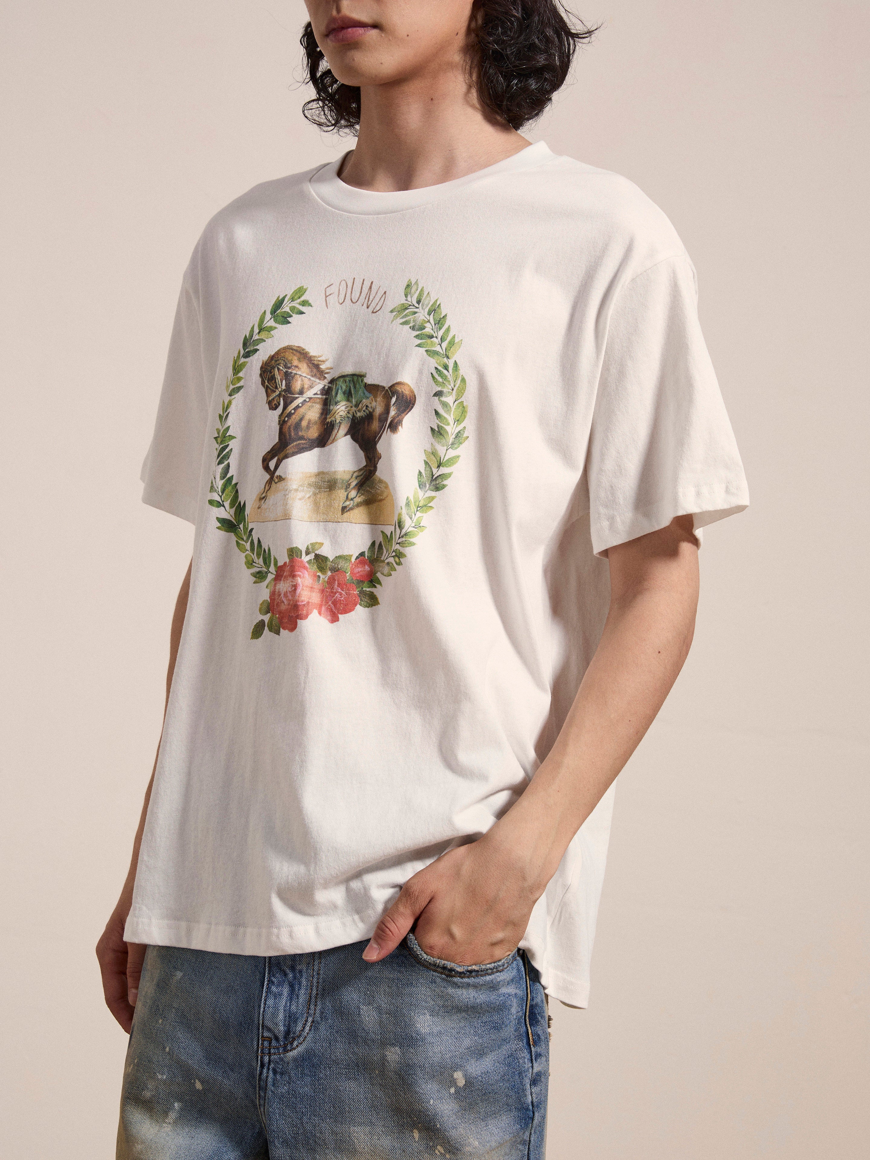 A person wearing a white FOUND Rodeo Tee featuring a horse and floral design, paired with blue jeans, with one hand in their pocket.